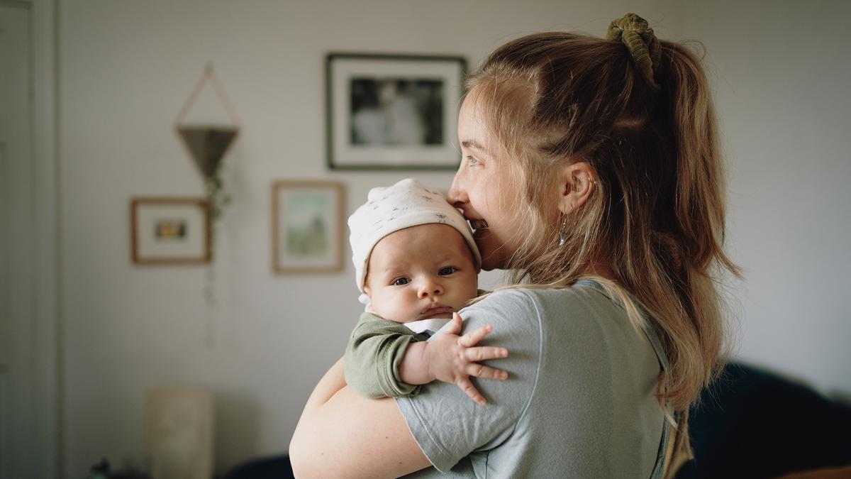 A woman holding a baby in a cosy living room, with framed pictures on the wall in the background.