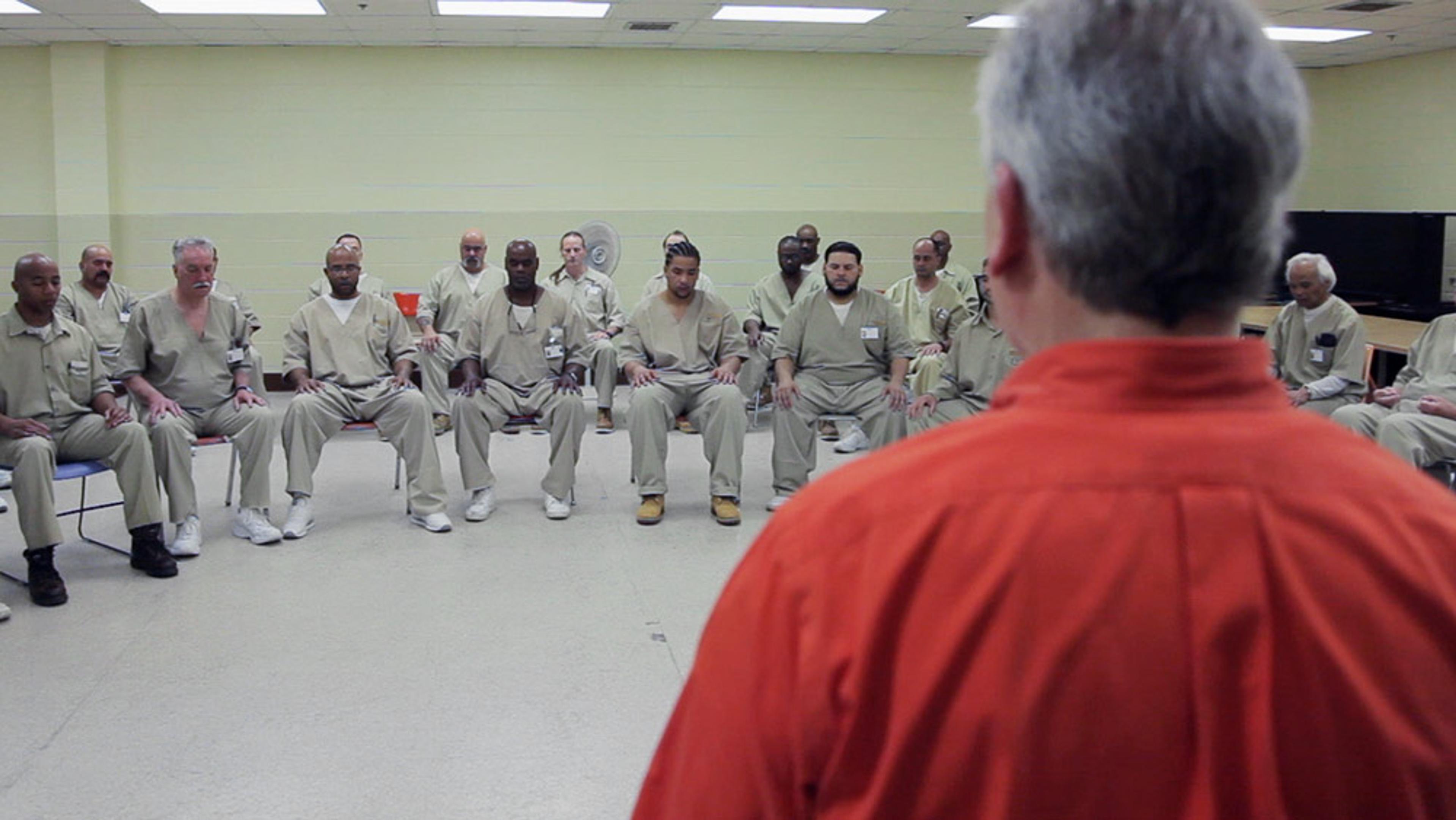 A group of men in prison uniforms sitting in a circle in a room, viewed from behind a man in an orange uniform.