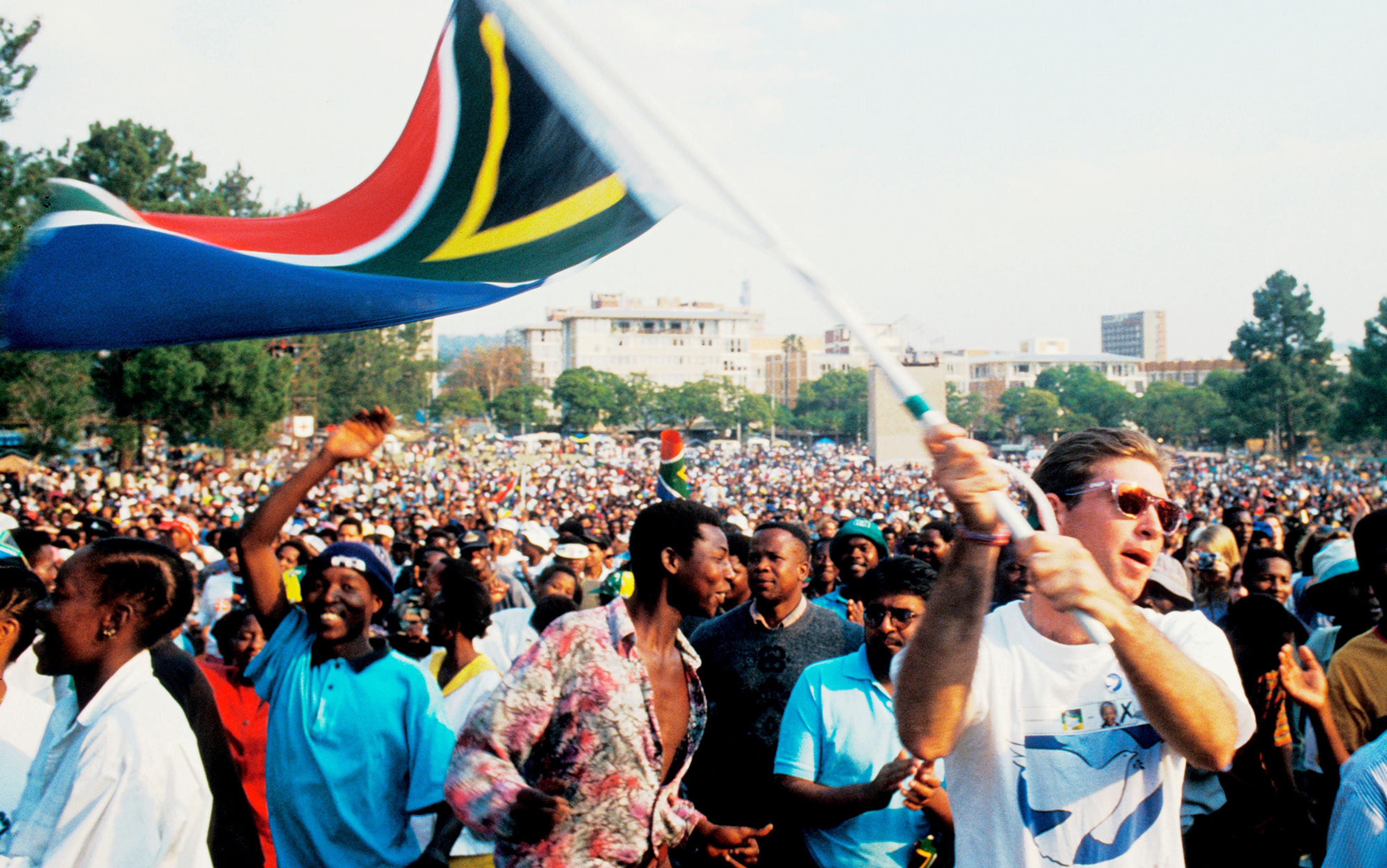 Photo of a large crowd celebrating outdoors with a person waving a South African flag energetically in the foreground.