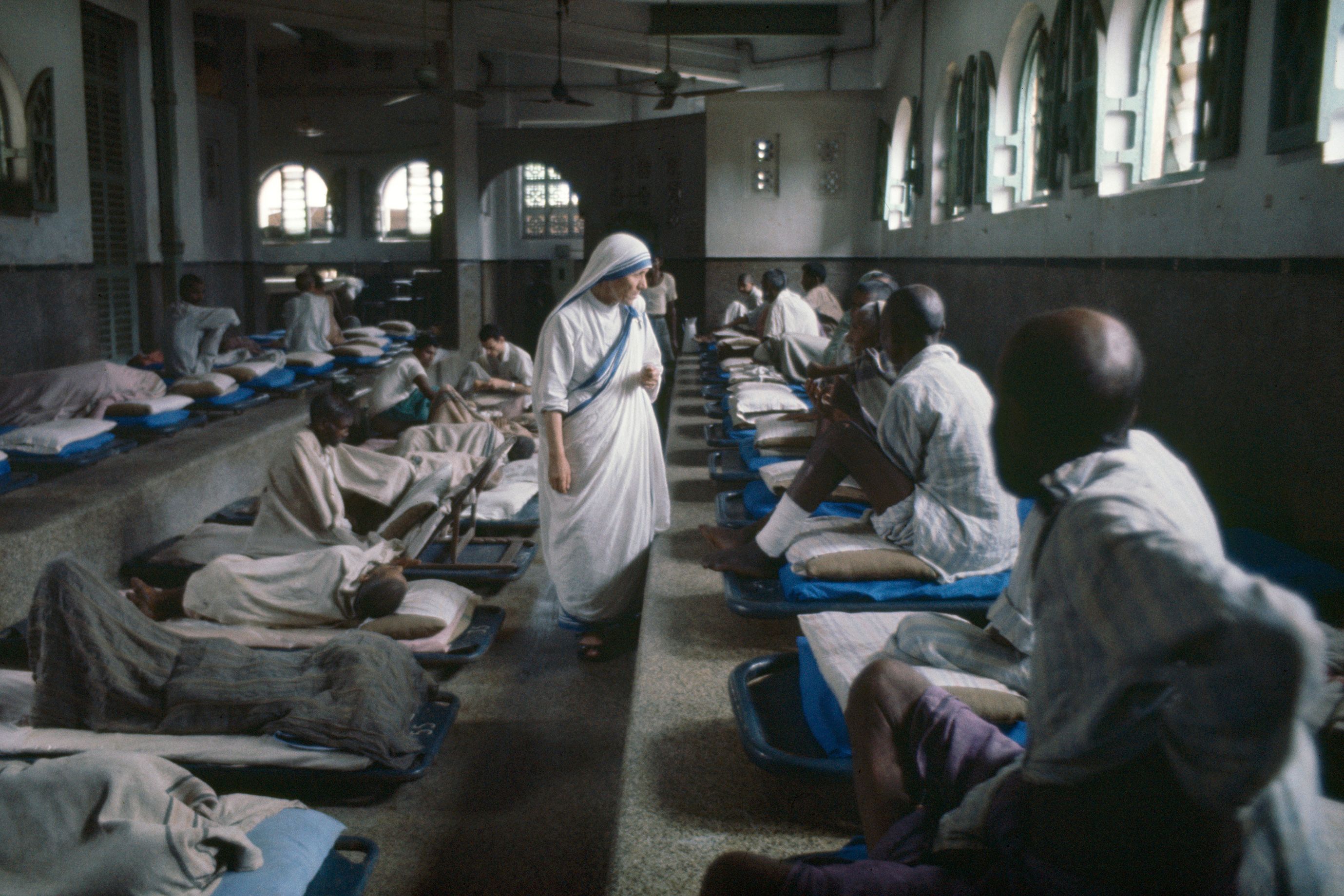 Photo of a nun in a hospital ward walking between patients lying on beds lined up against the walls, with light from windows.
