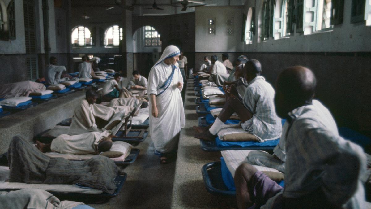 Photo of a nun in a hospital ward walking between patients lying on beds lined up against the walls, with light from windows.