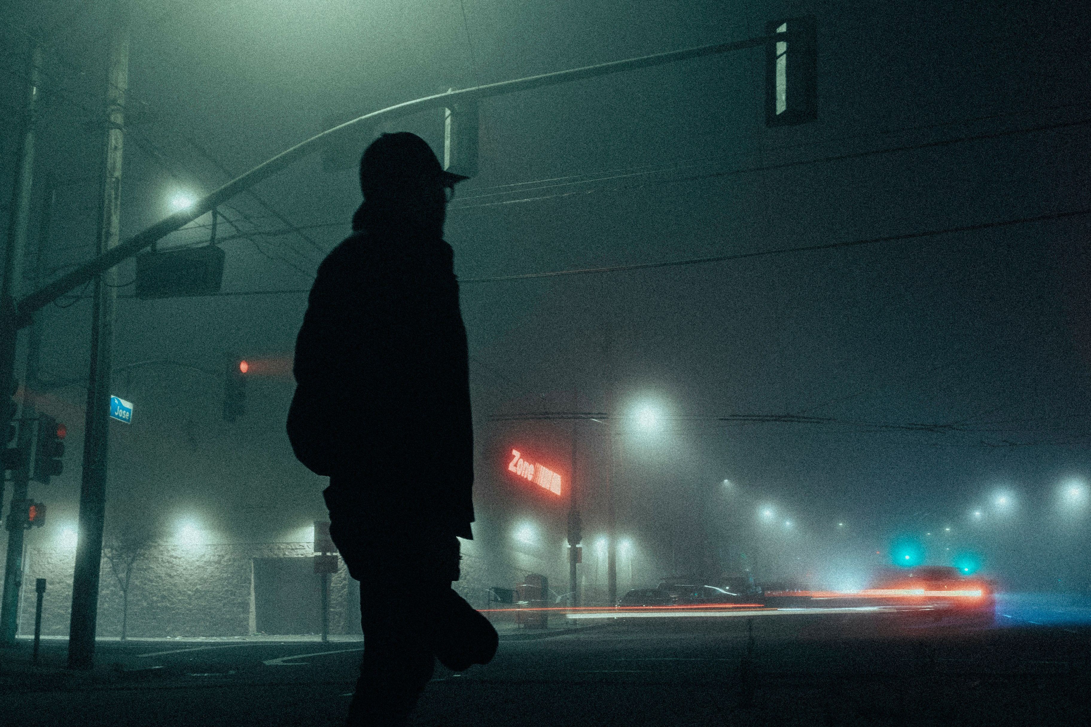 A silhouetted figure on a foggy street corner with traffic lights and blurred car lights at night.