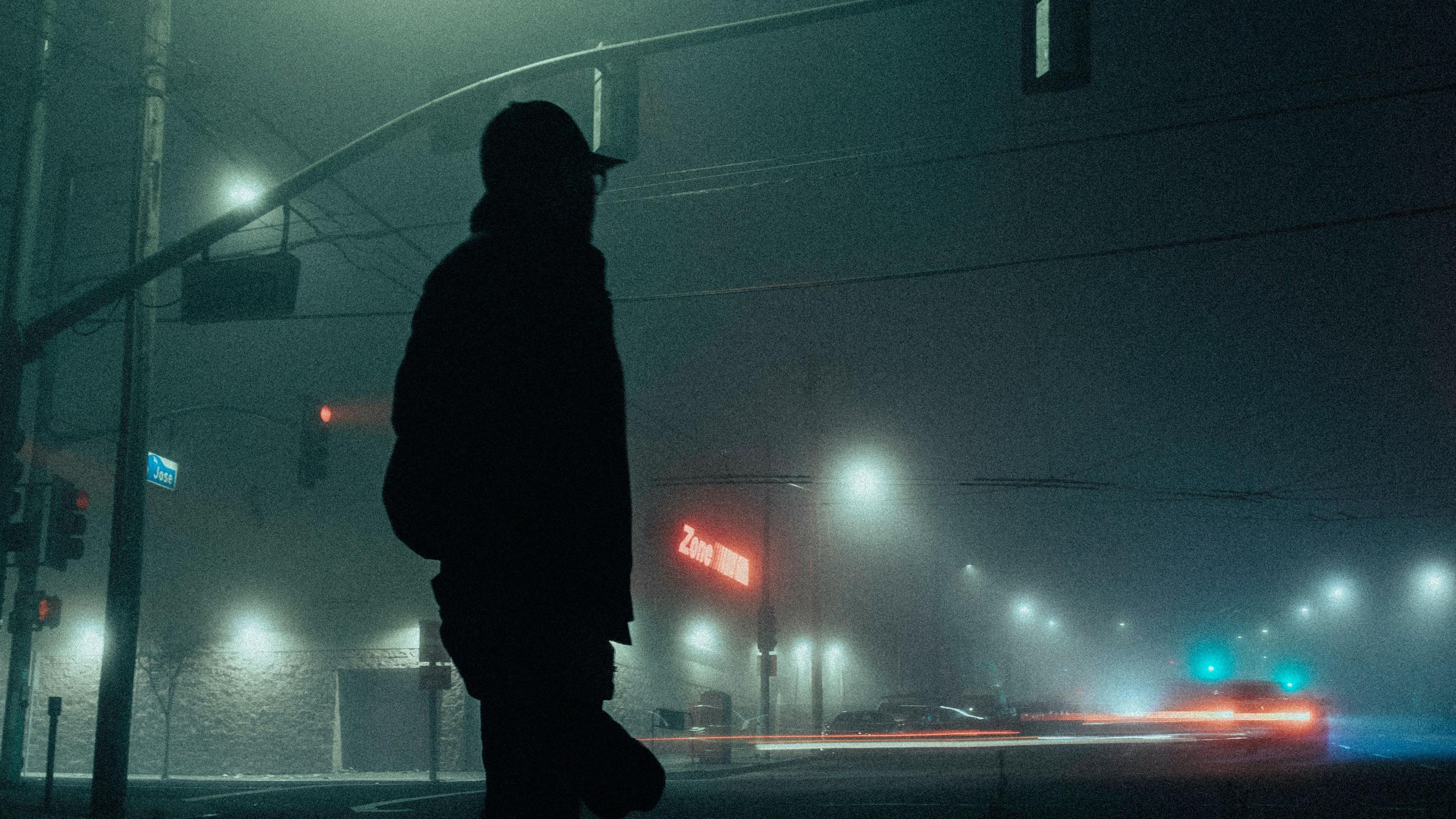 A silhouetted figure on a foggy street corner with traffic lights and blurred car lights at night.