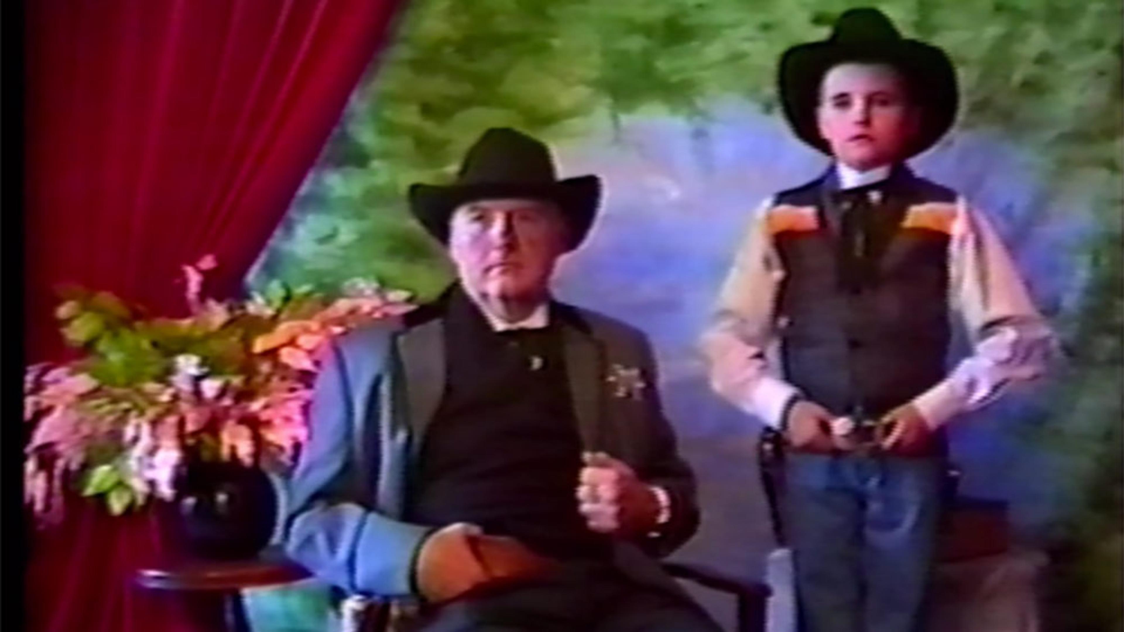 A vintage photo of a man and boy in cowboy attire. The man sits by a table with flowers, the boy stands with a hand on his belt.