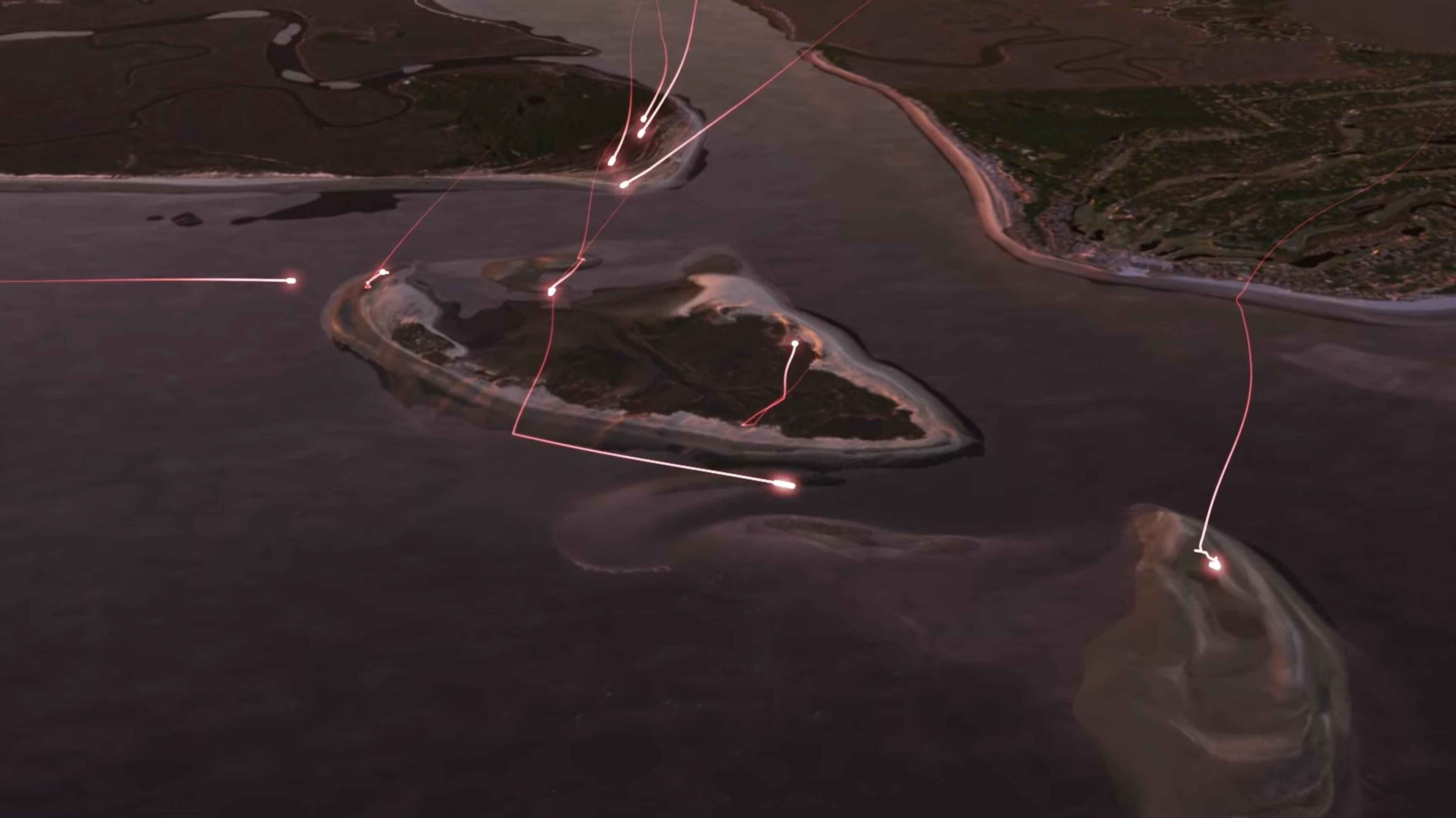 Aerial view of a coastal area at dusk with multiple streaks of red light travelling over water and land, illuminating the landscape.