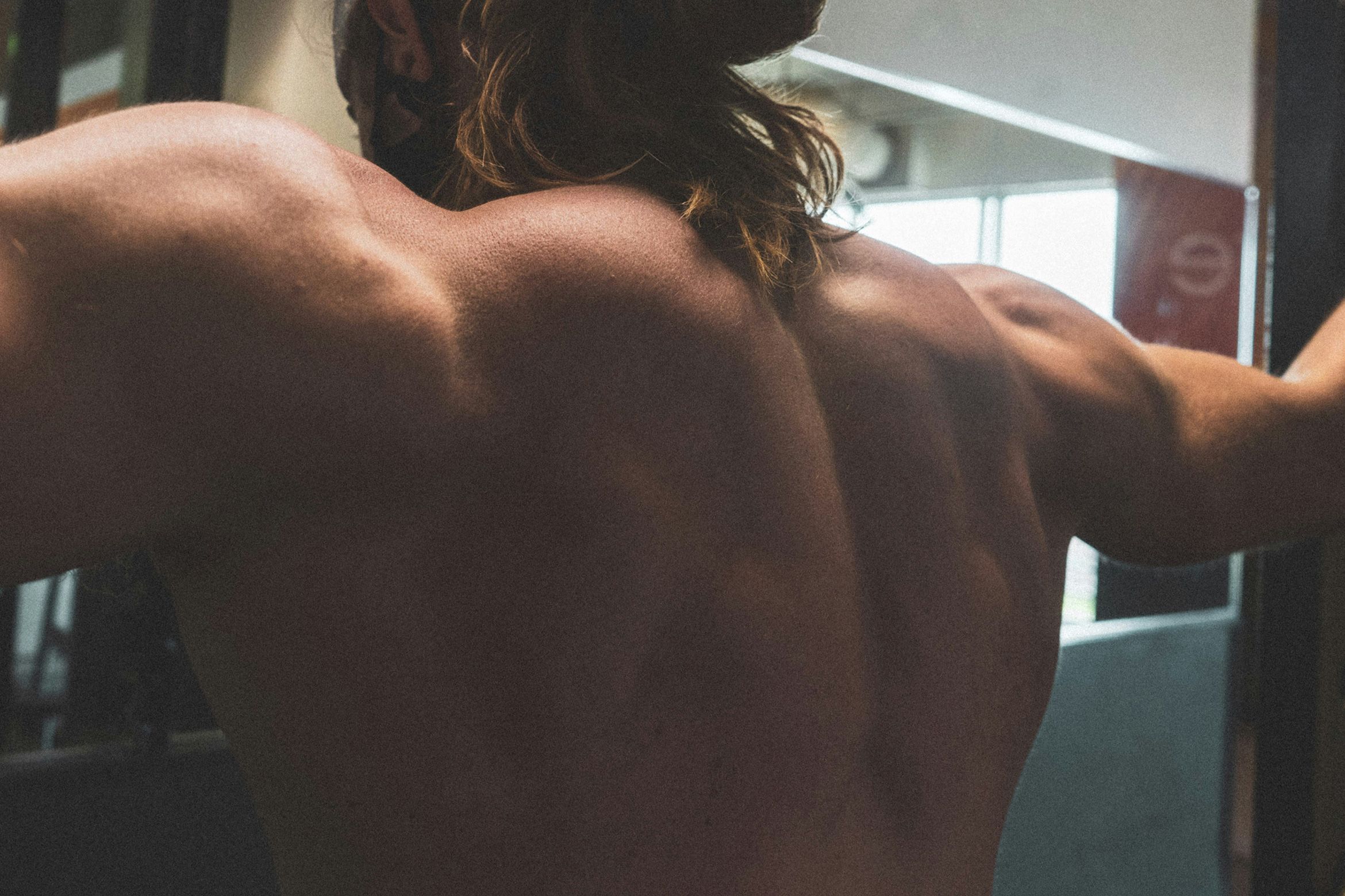A muscular man’s back and shoulders in a gym setting with long hair tied back.