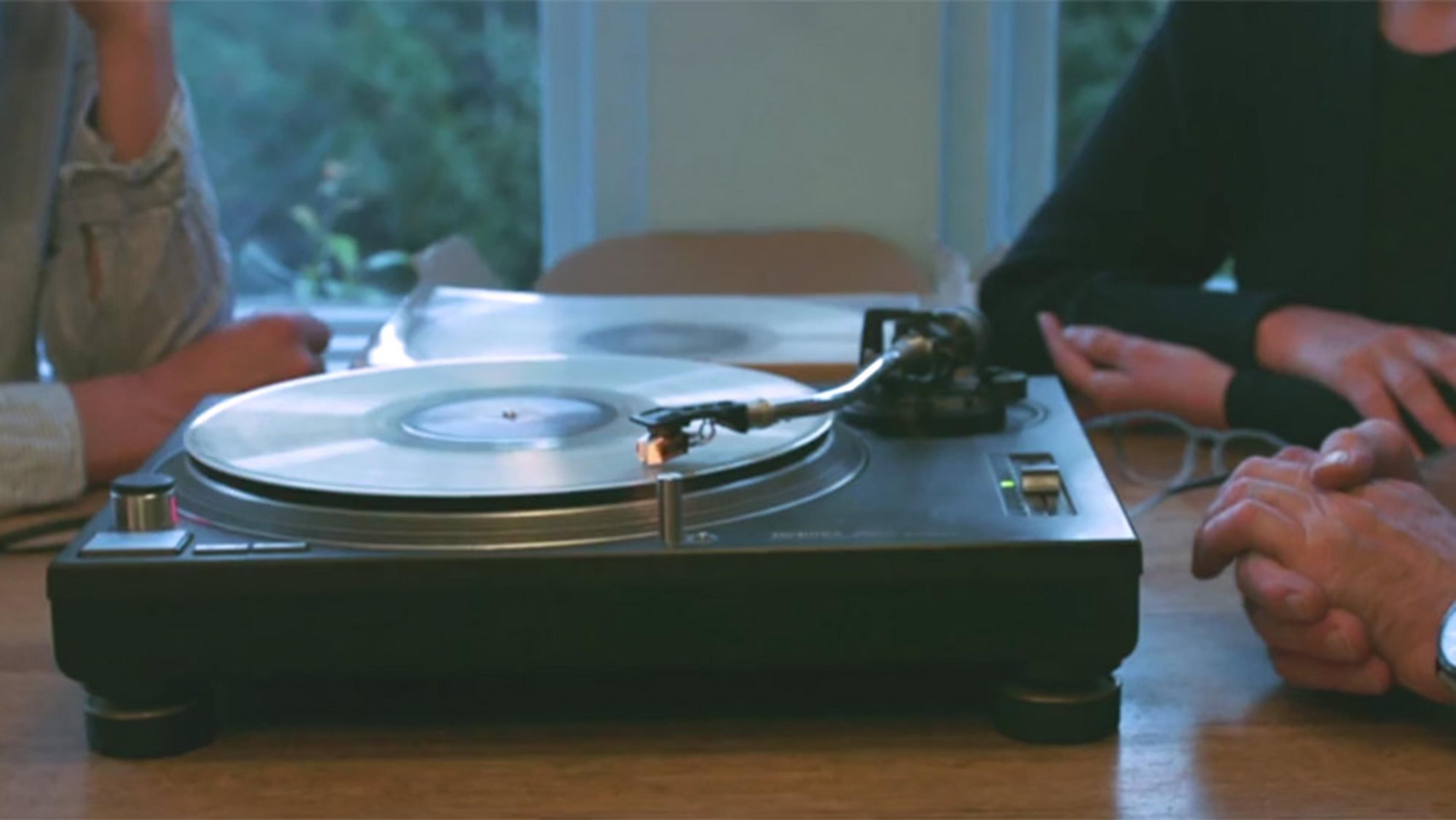 A turntable playing a vinyl record on a wooden table with people sitting around it, partially visible, in a cosy indoor setting.