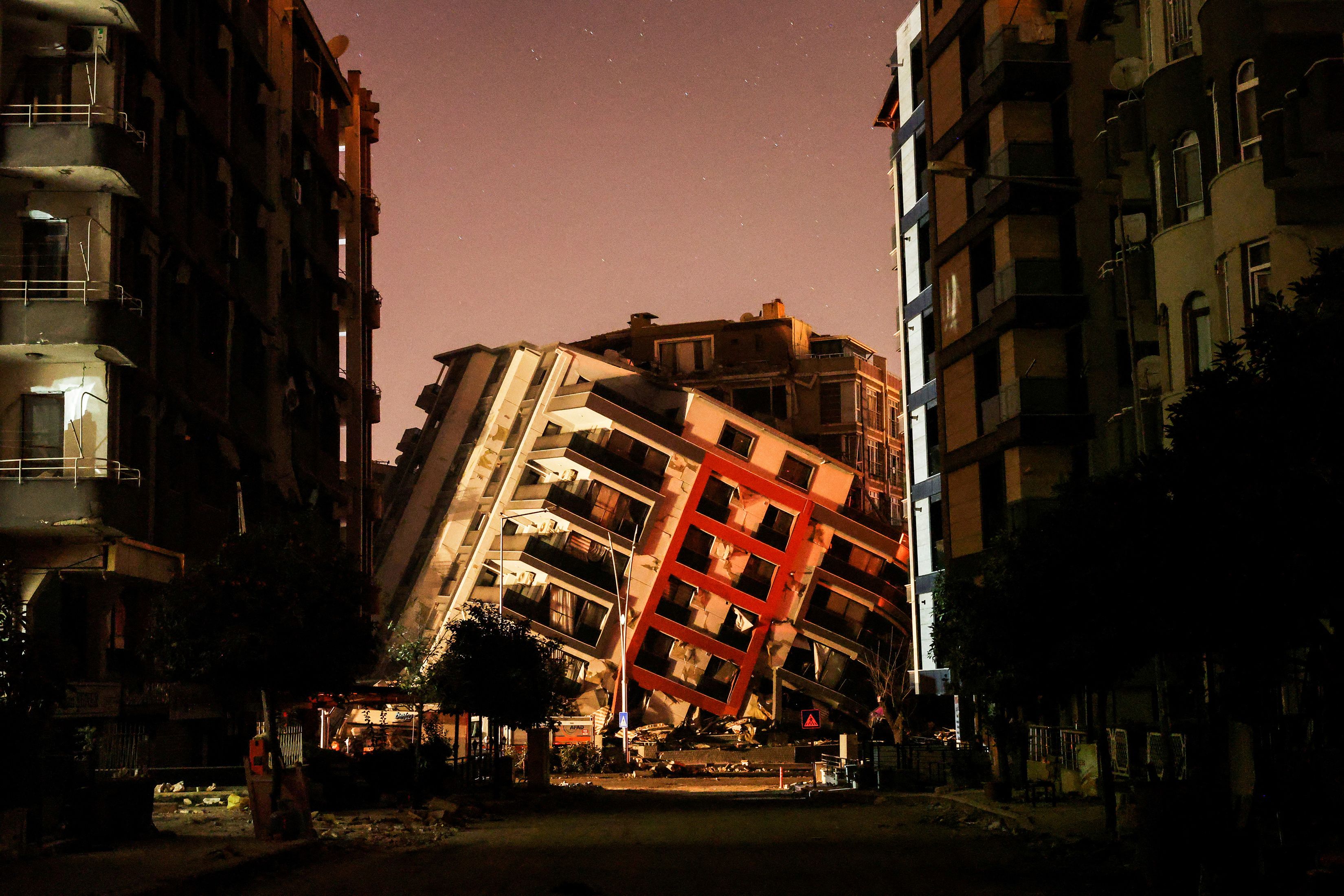 A collapsed apartment building leaning between other damaged buildings in twilight