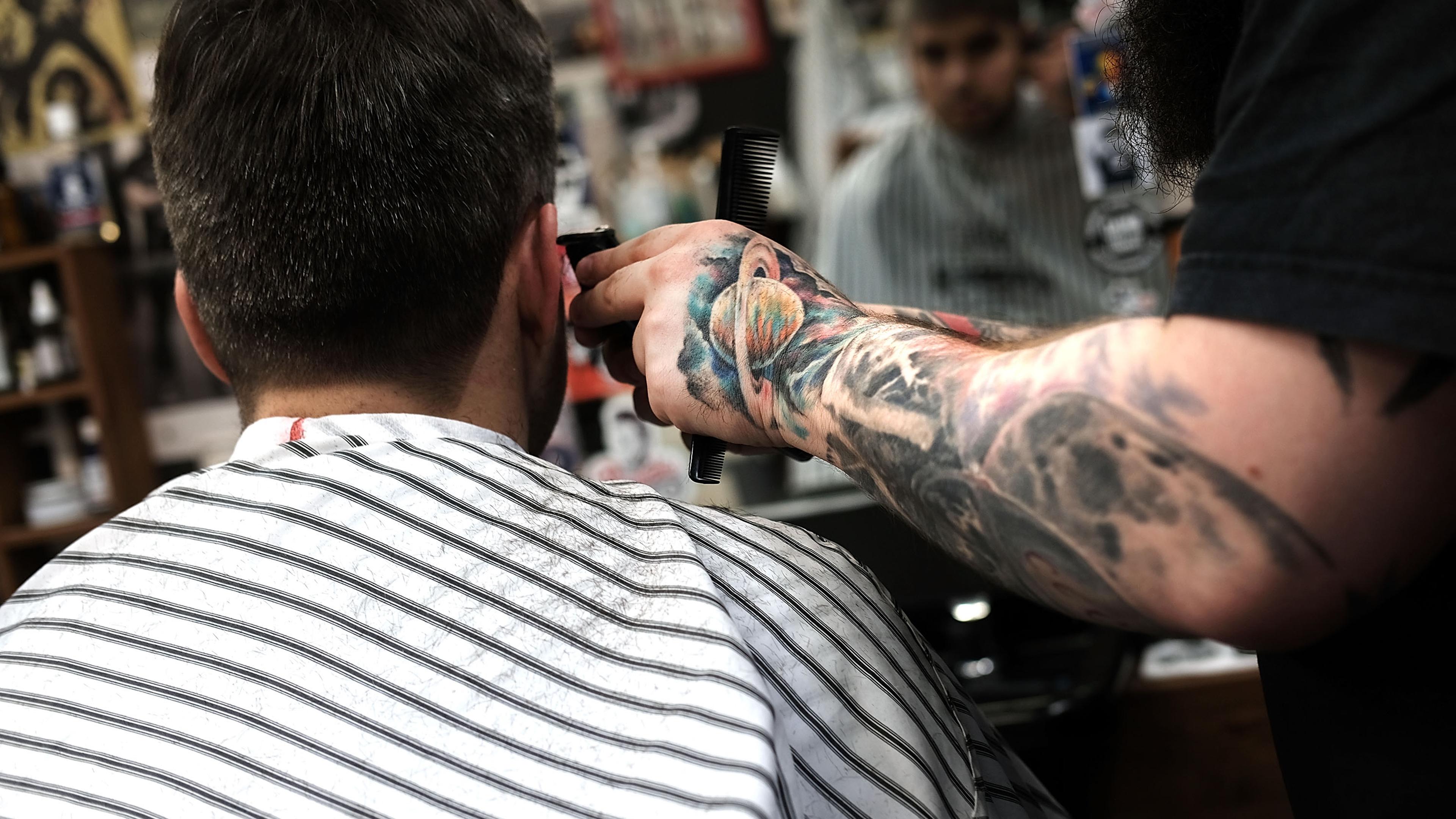Photo of a tattooed barber trimming a man’s hair in a striped cape, with a reflection in the mirror.