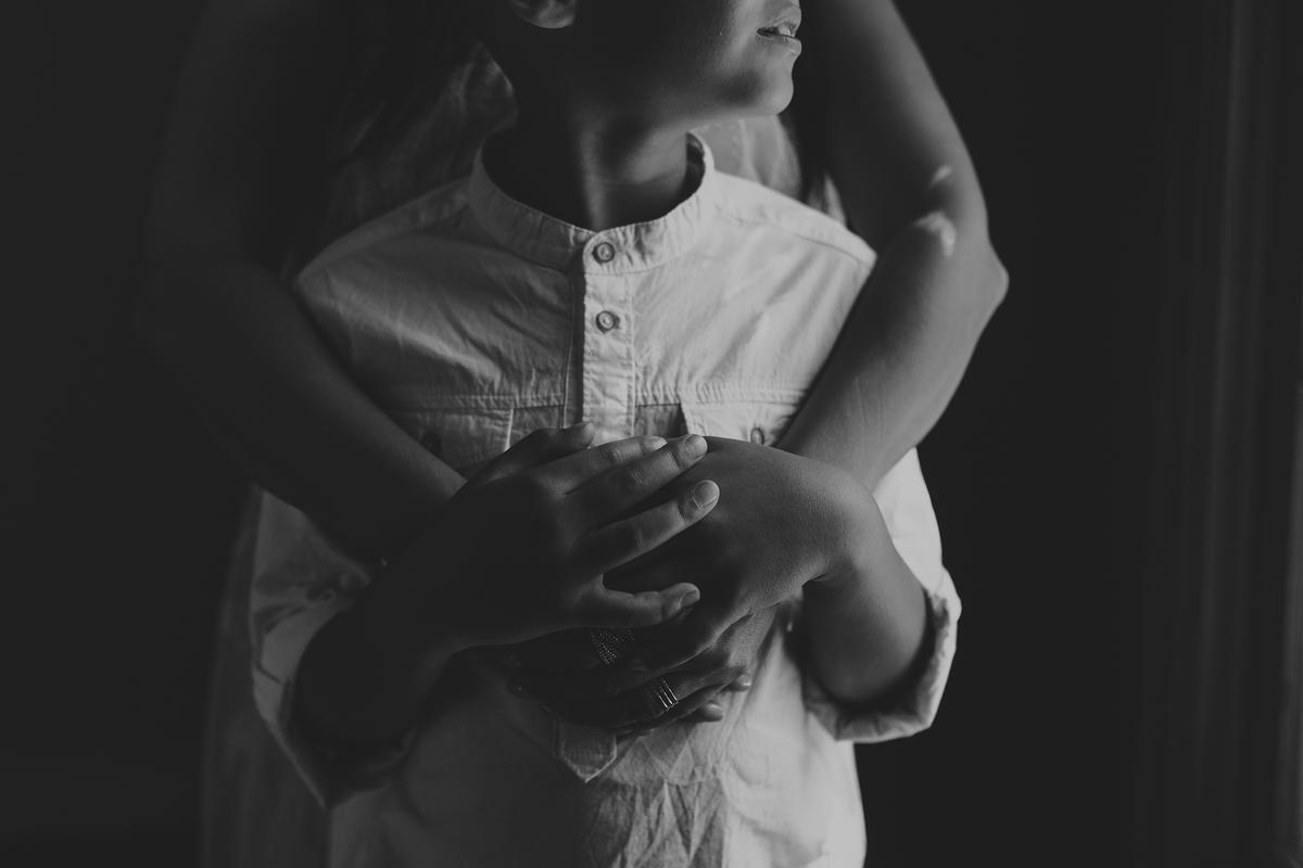 Black and white photo of a child in a white shirt embraced from behind by an adult, only their hands and torso are visible.
