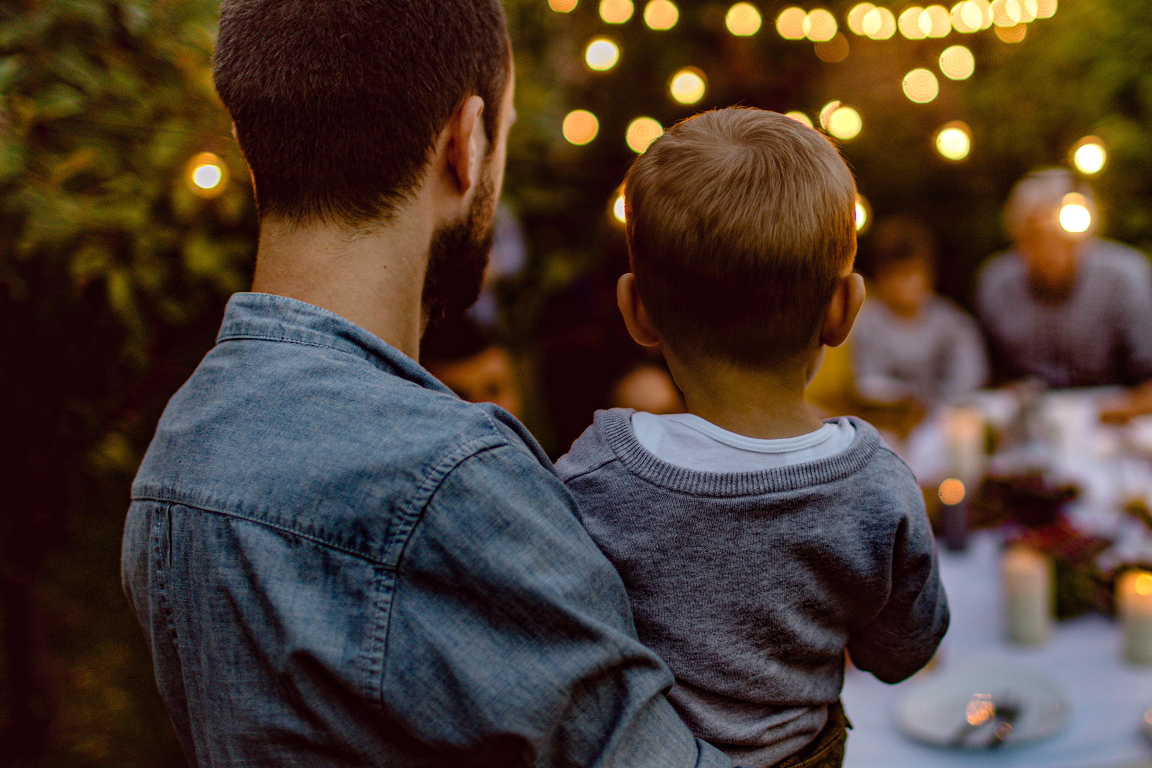 Photo of a man holding a child outdoors at a warmly lit gathering, string lights in the background.