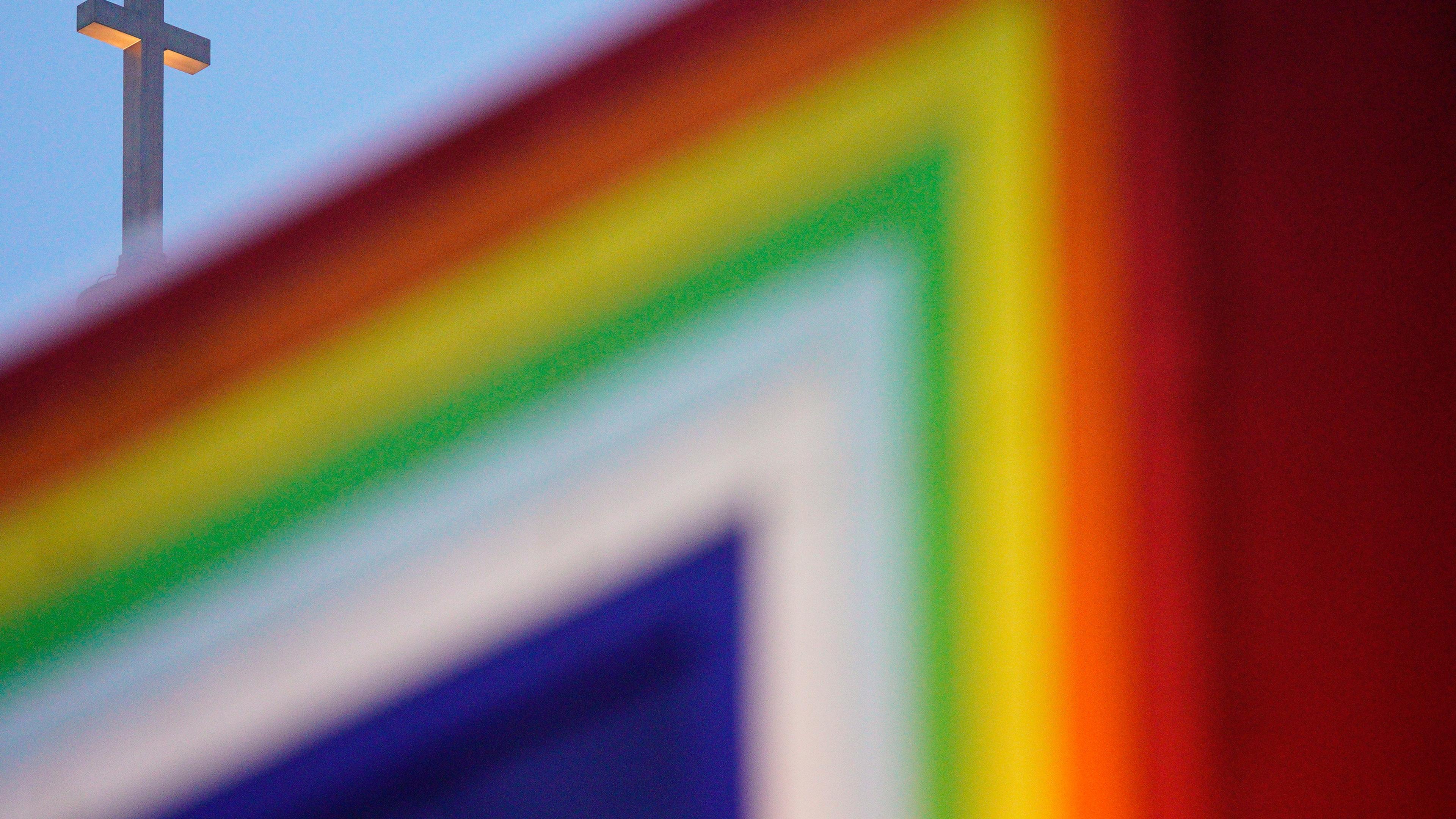 Photo of a cross in the sky with a blurred rainbow-coloured flag in the foreground.
