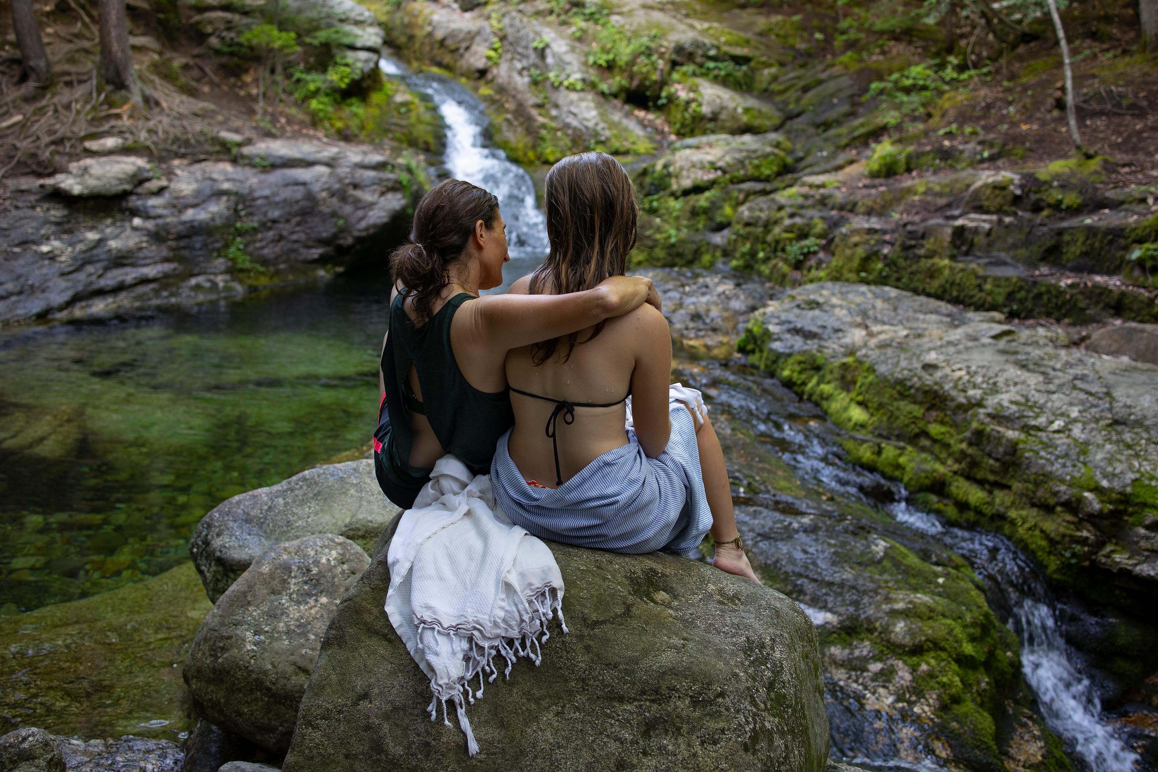 Photo of two people sitting on a rock by a stream surrounded by greenery, one with an arm around the other.