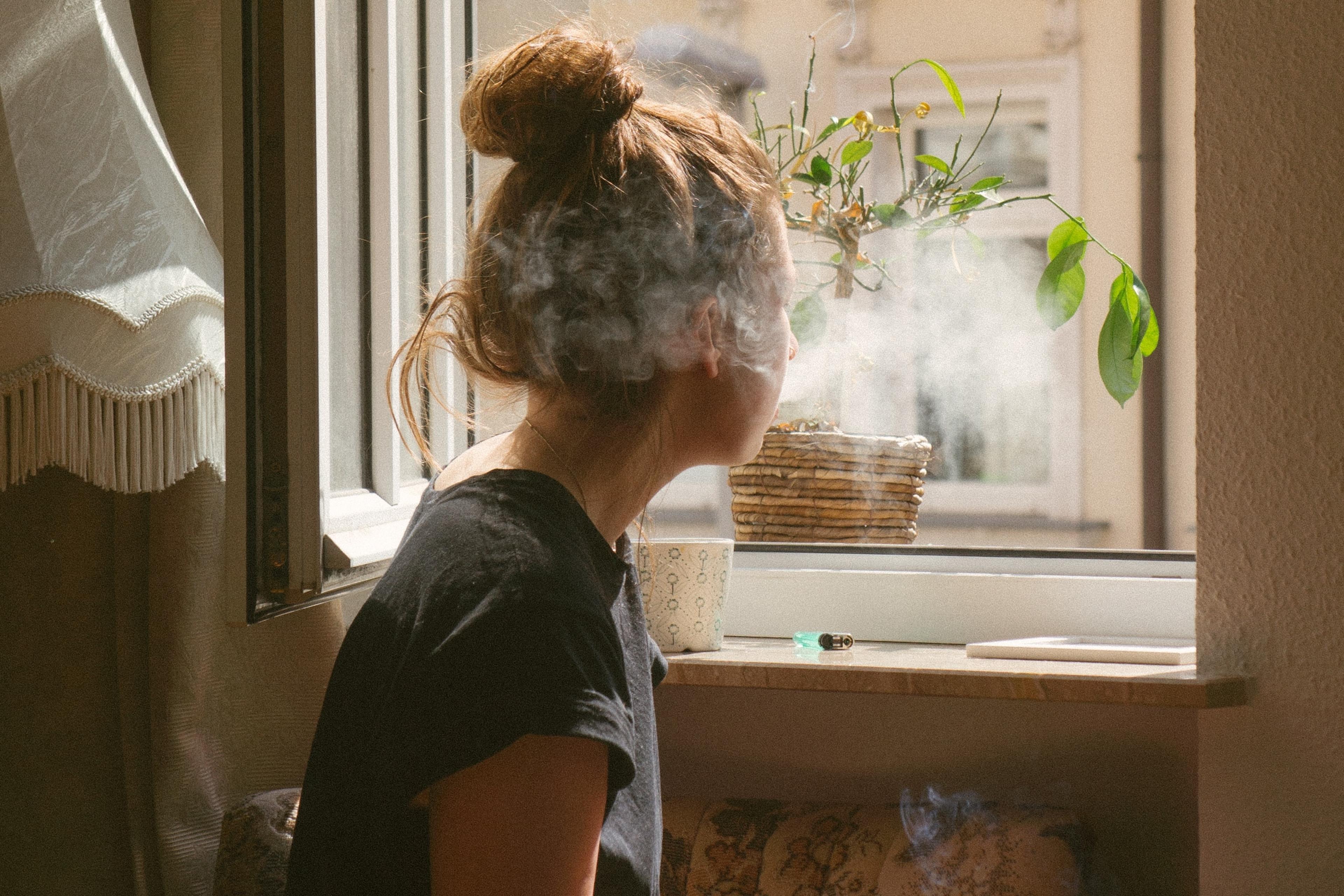 Photo of a person with a bun sitting by a window smoking. Sunlight streams in highlighting smoke tendrils and a plant.
