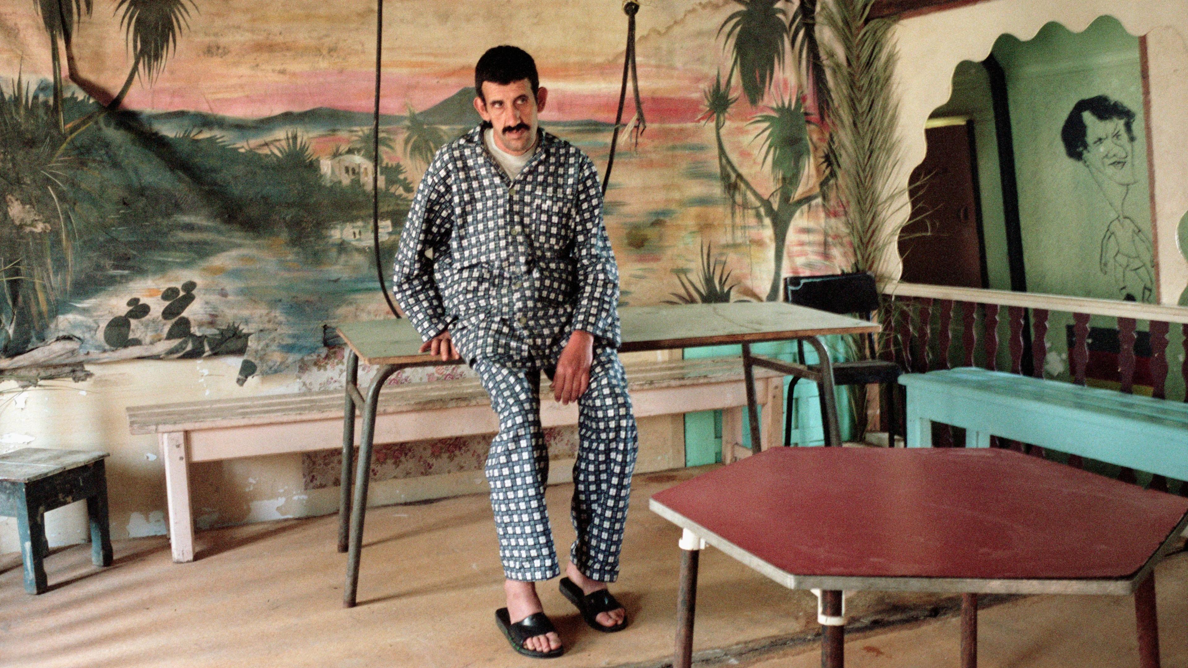 A photo of a man in patterned clothing sitting on tables in a room with mural of palm trees and water, surrounded by furniture.