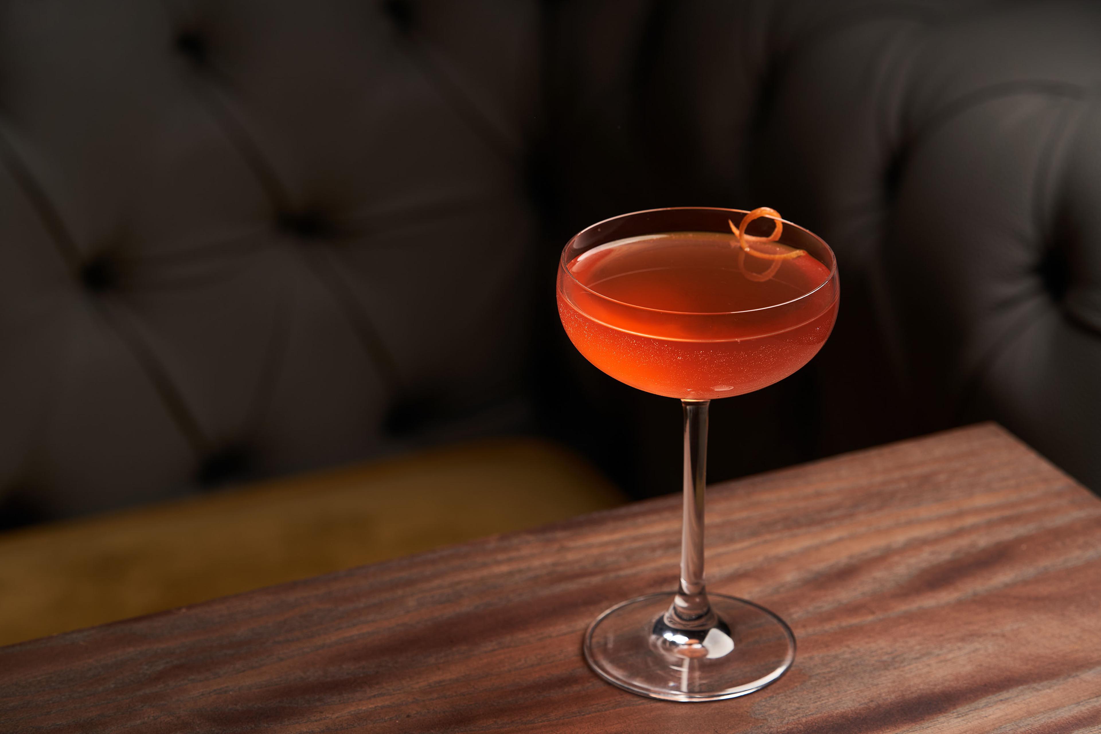 A cocktail in a coupe glass with an orange twist on a wooden table in front of a dark, tufted lounge seating