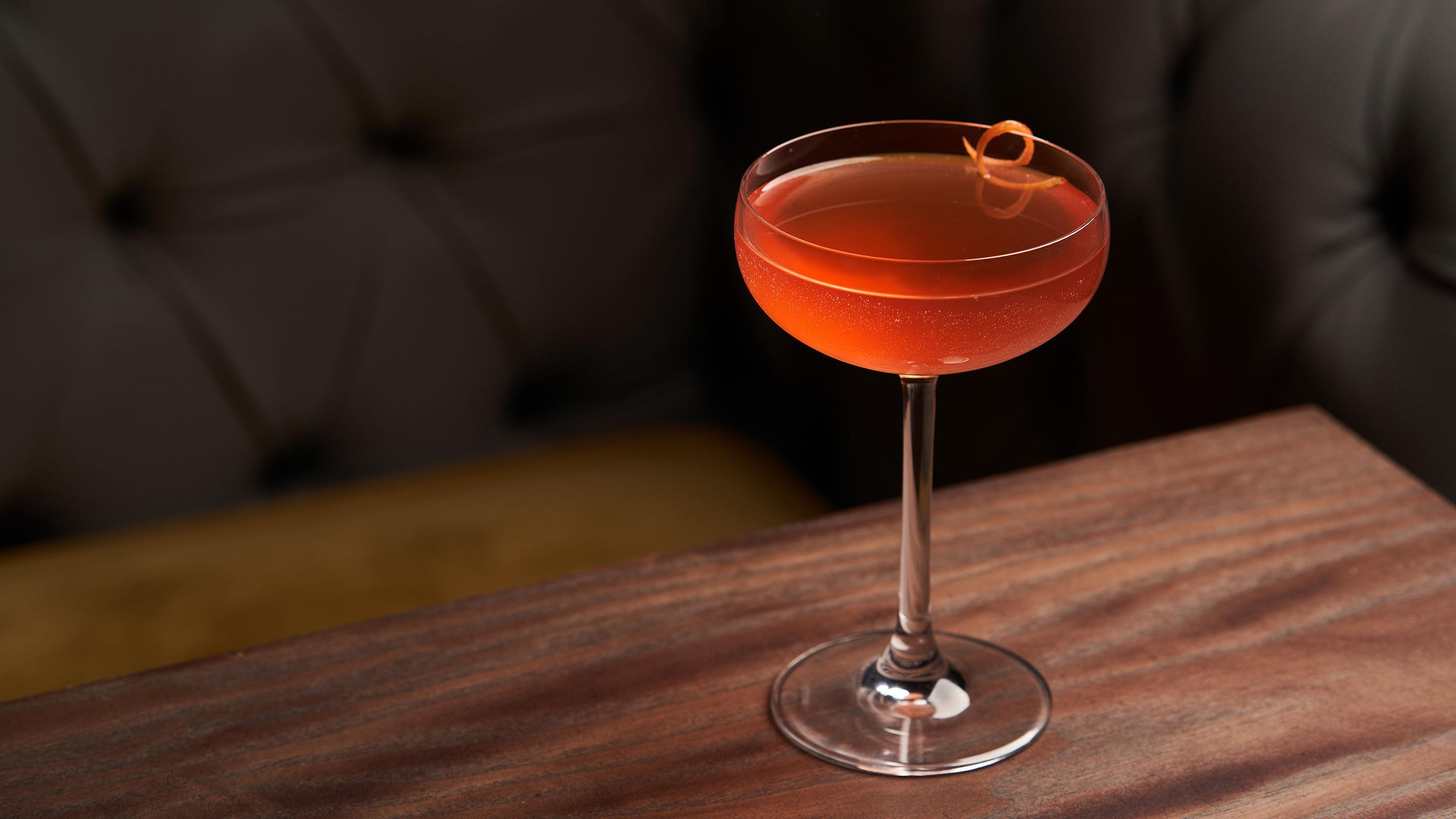 A cocktail in a coupe glass with an orange twist on a wooden table in front of a dark, tufted lounge seating