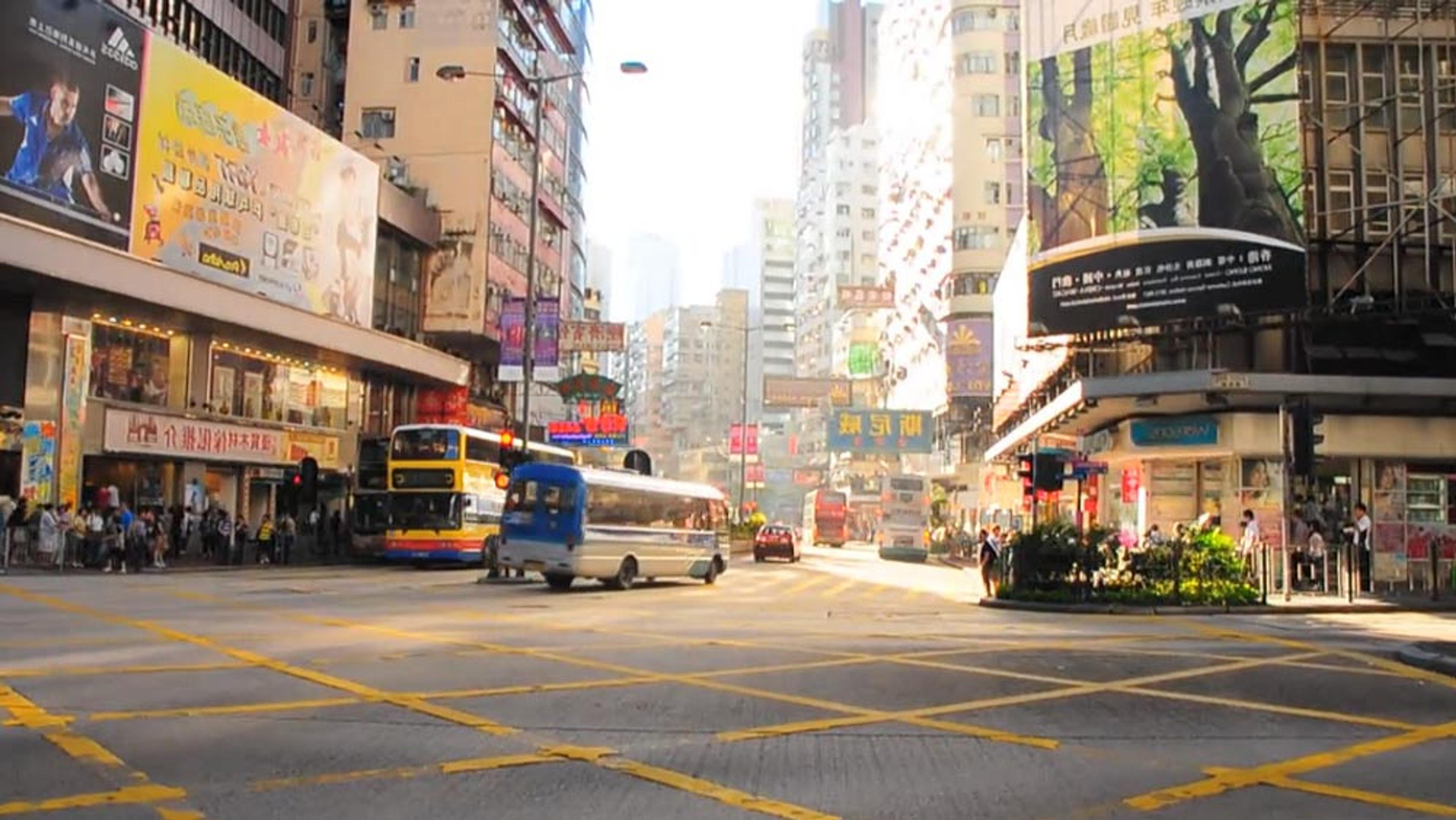 A busy city intersection with buses, cars and pedestrians surrounded by tall buildings and large billboards in daytime.