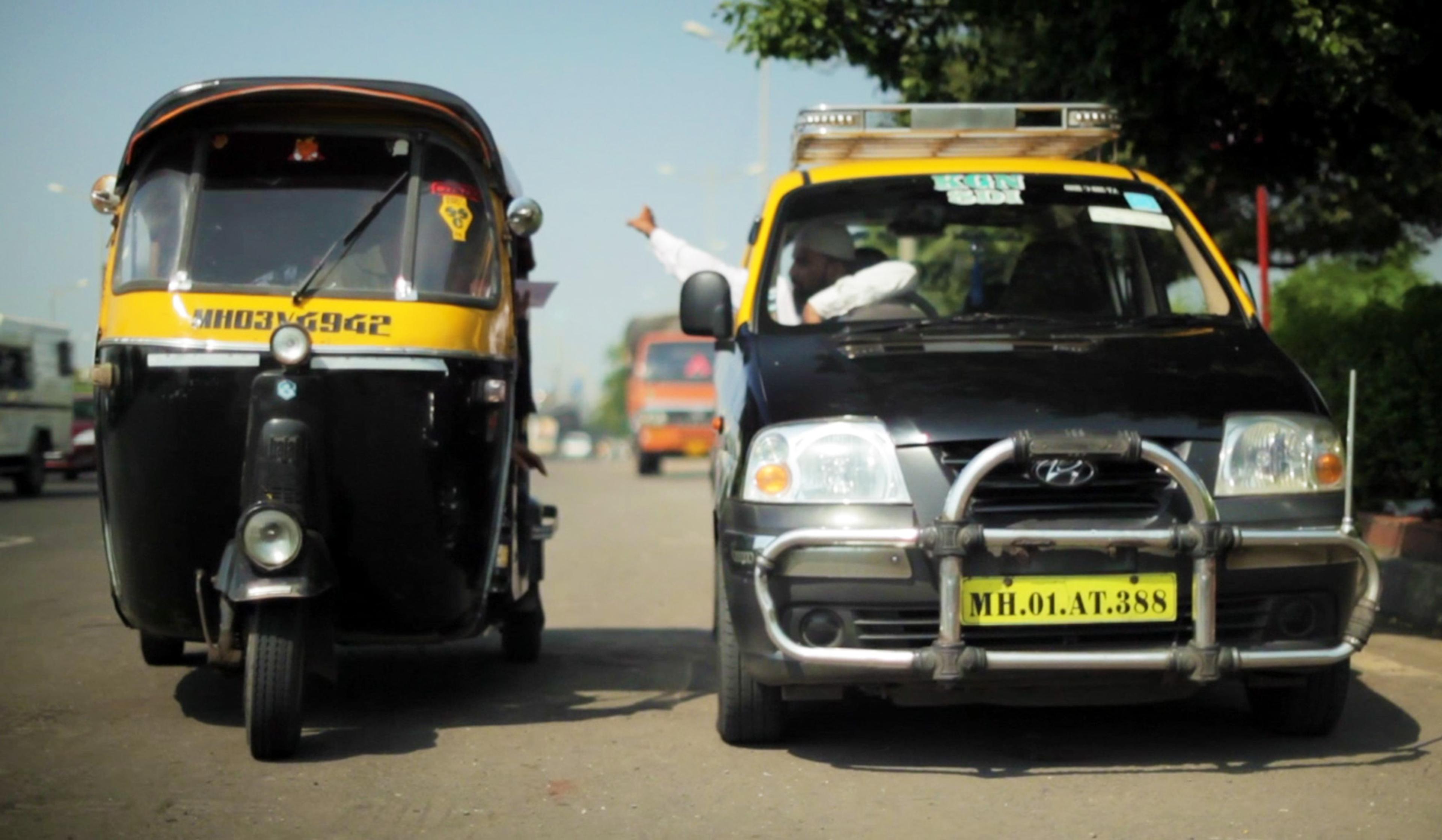 Auto rickshaw and black taxi on a road, with drivers conversing through the windows under a clear sky.