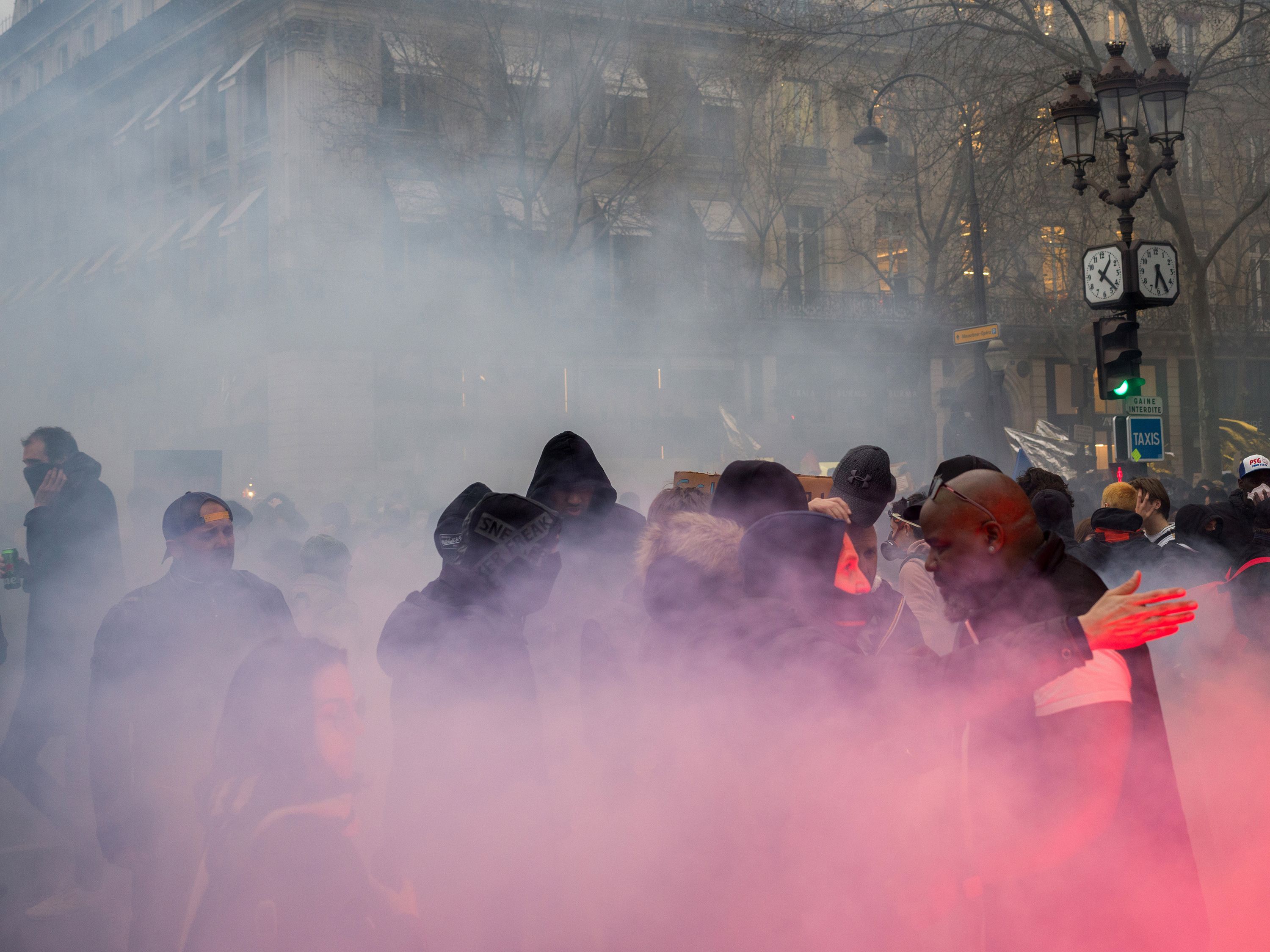 A crowd enveloped in pink smoke on a city street, with people wearing hoodies and jackets, and buildings in the background.