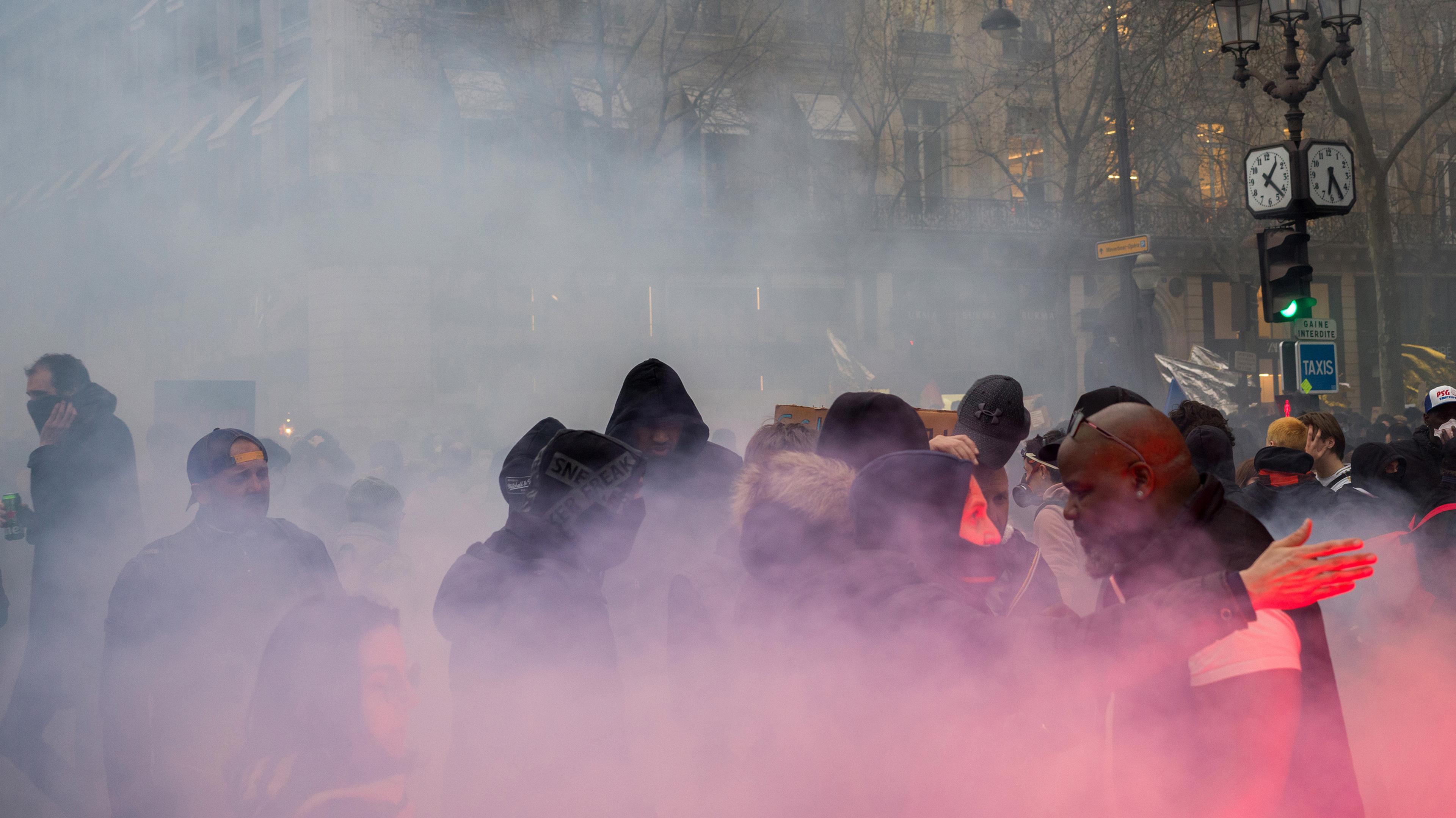 A crowd enveloped in pink smoke on a city street, with people wearing hoodies and jackets, and buildings in the background.