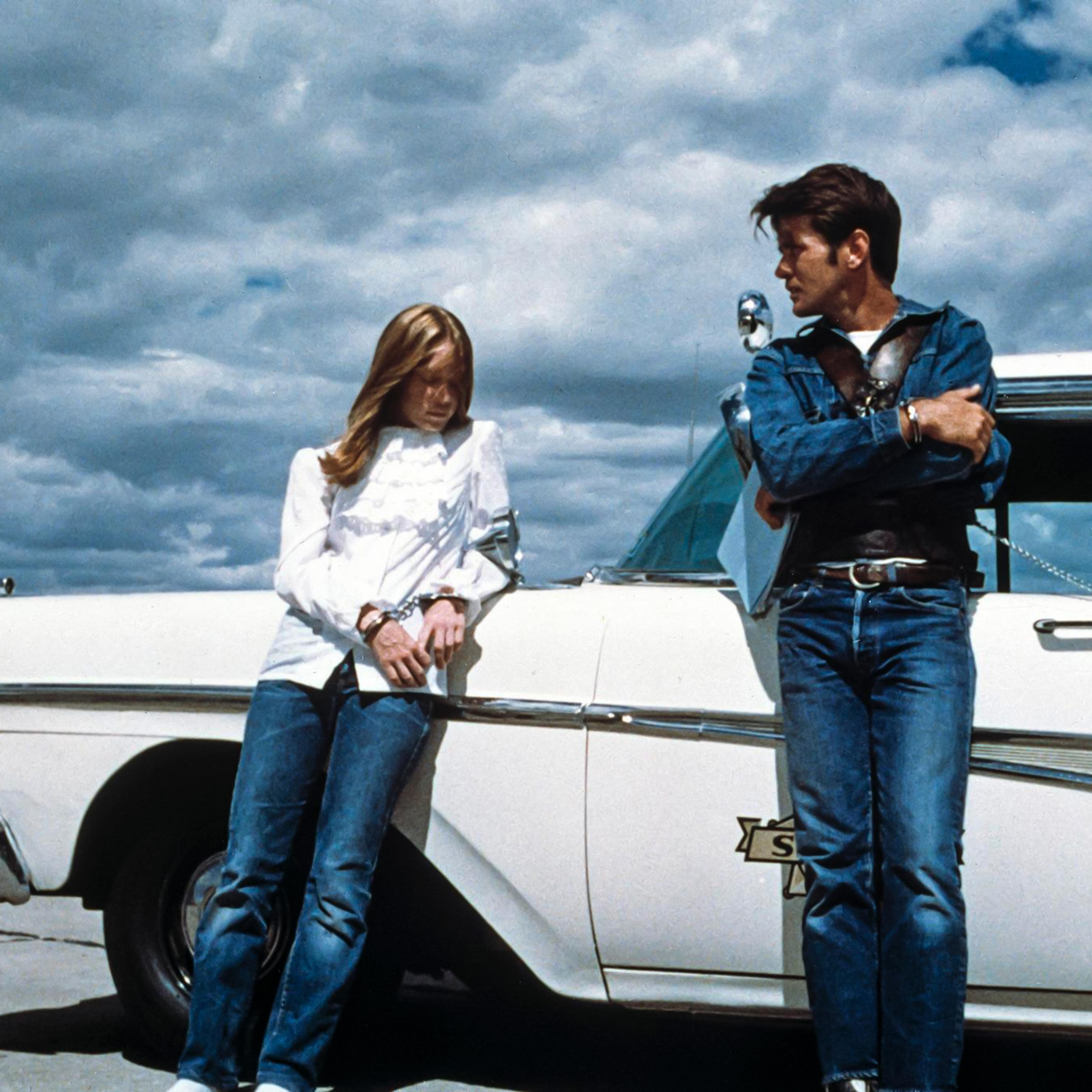 A young couple in handcuffs lean against a 1950s police car beneath a brooding prairie sky