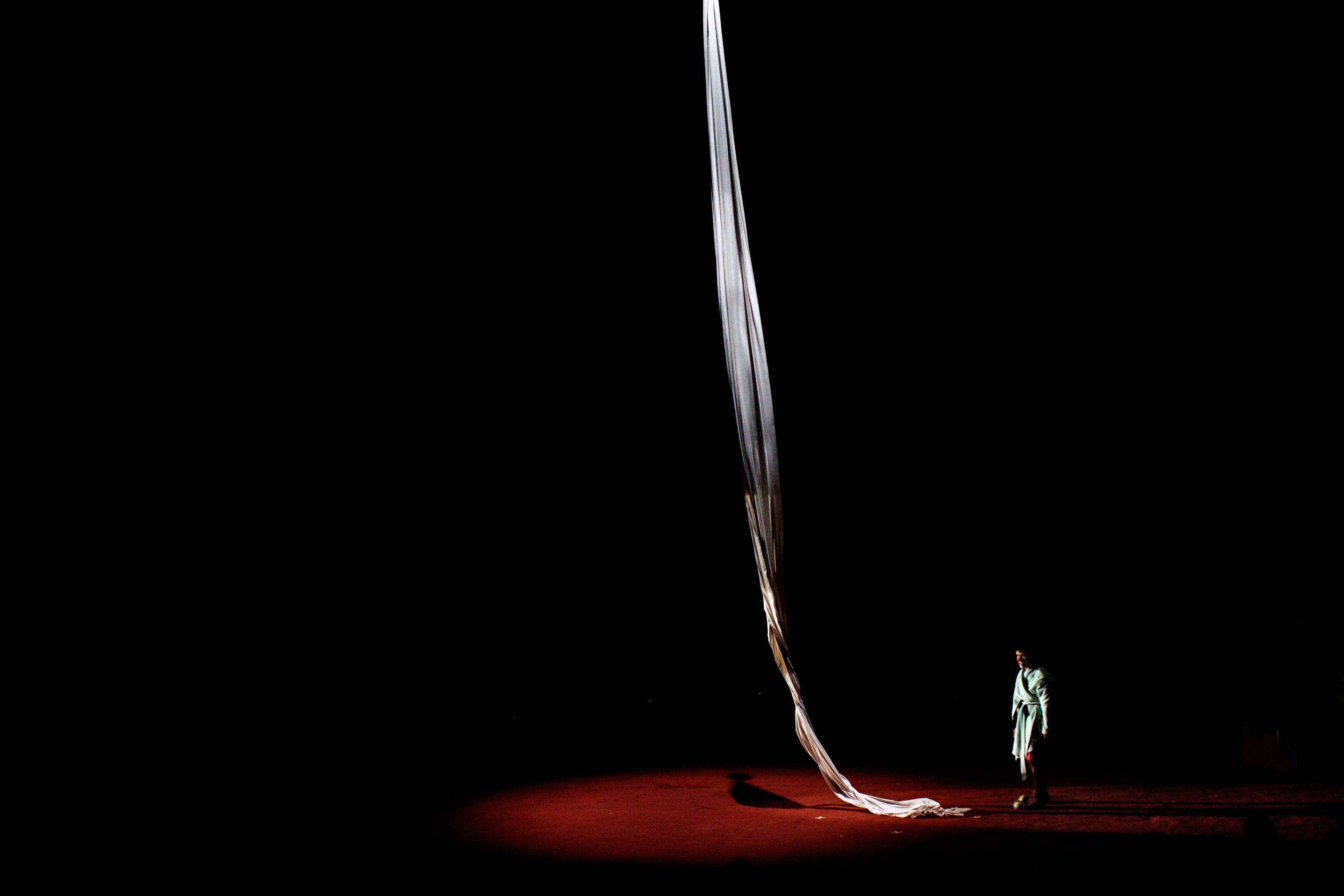 A person stands under a spotlight next to a long flowing fabric on a dark stage; photo.