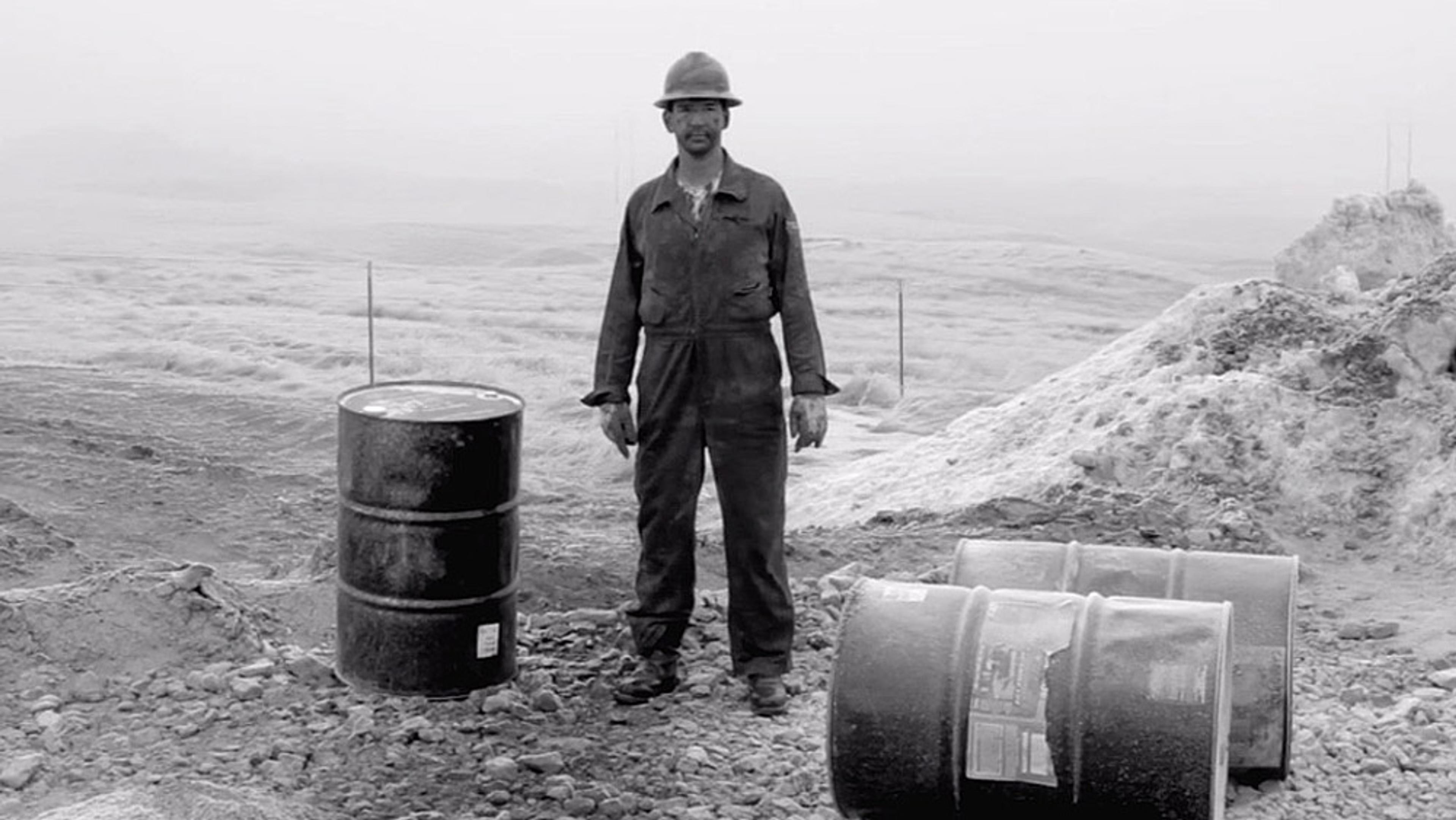 Black and white photo of a man in work clothes and helmet standing outdoors beside a barrel in a barren, rocky landscape.