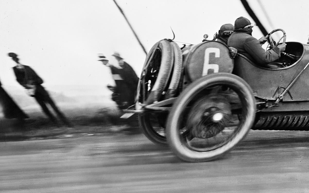 Black and white photo of a vintage race car numbered 6 speeding past blurred spectators emphasising motion and early motor racing.