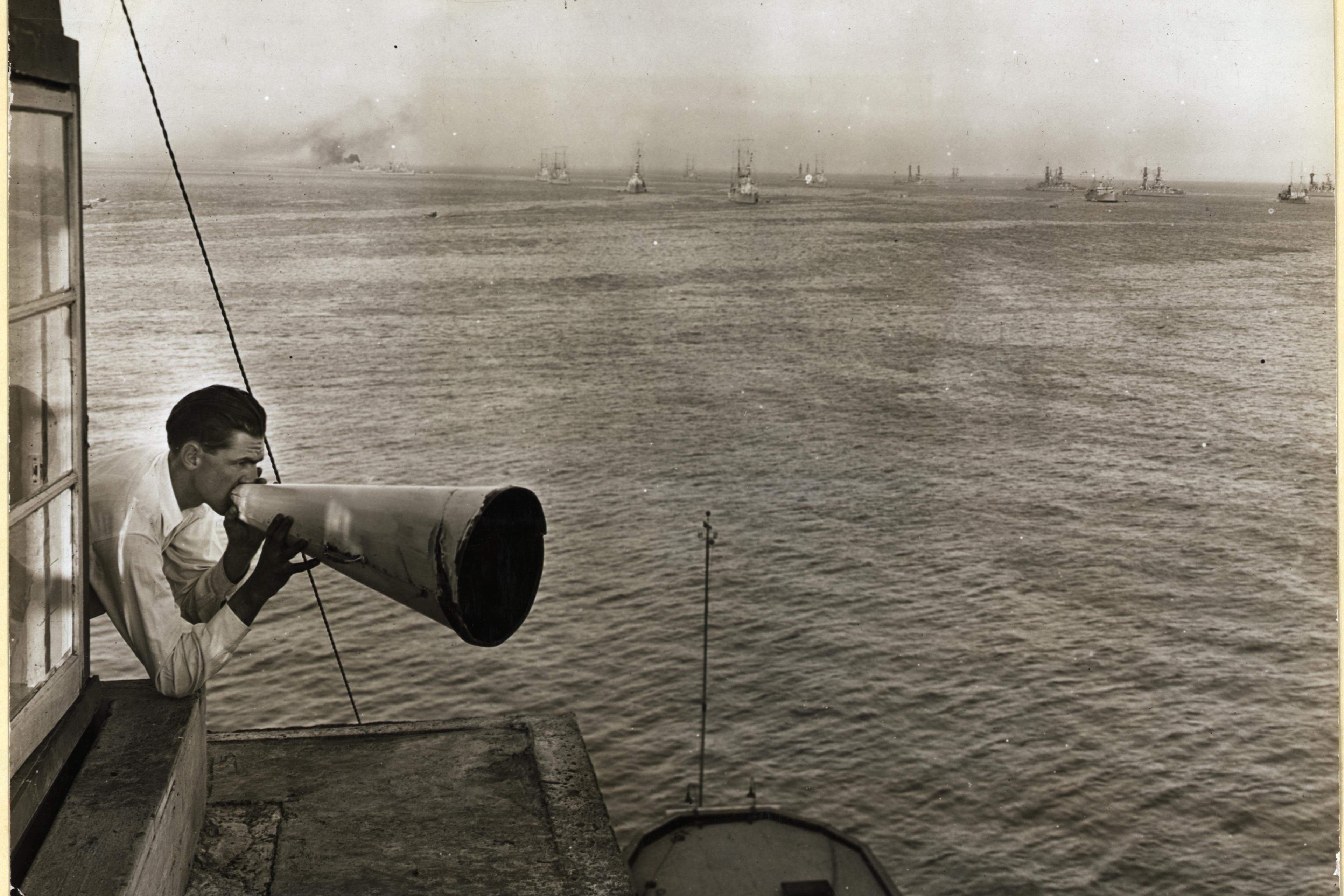 A black-and-white photo of a man using a megaphone over the sea with ships in the distance.