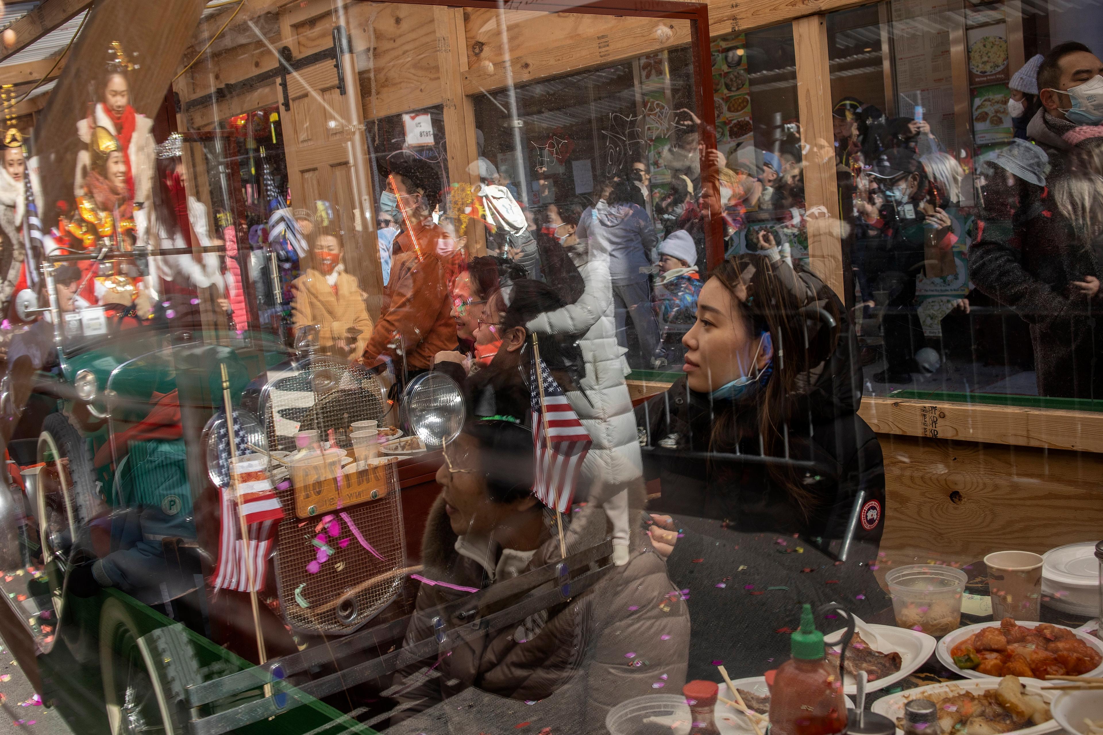 Photo of a crowded street scene reflecting in a window while people eat inside a restaurant, with flags and confetti visible.