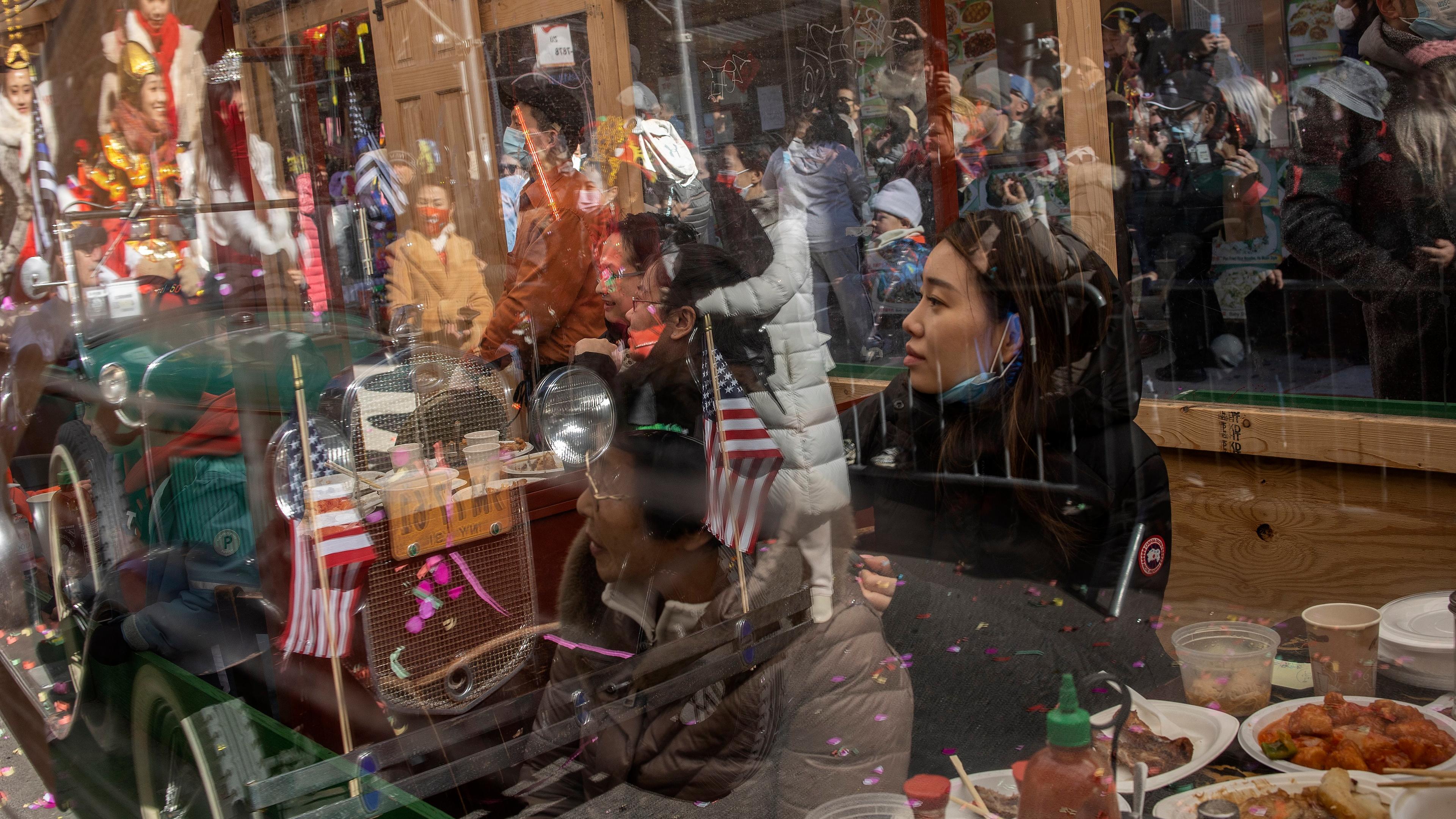 Photo of a crowded street scene reflecting in a window while people eat inside a restaurant, with flags and confetti visible.