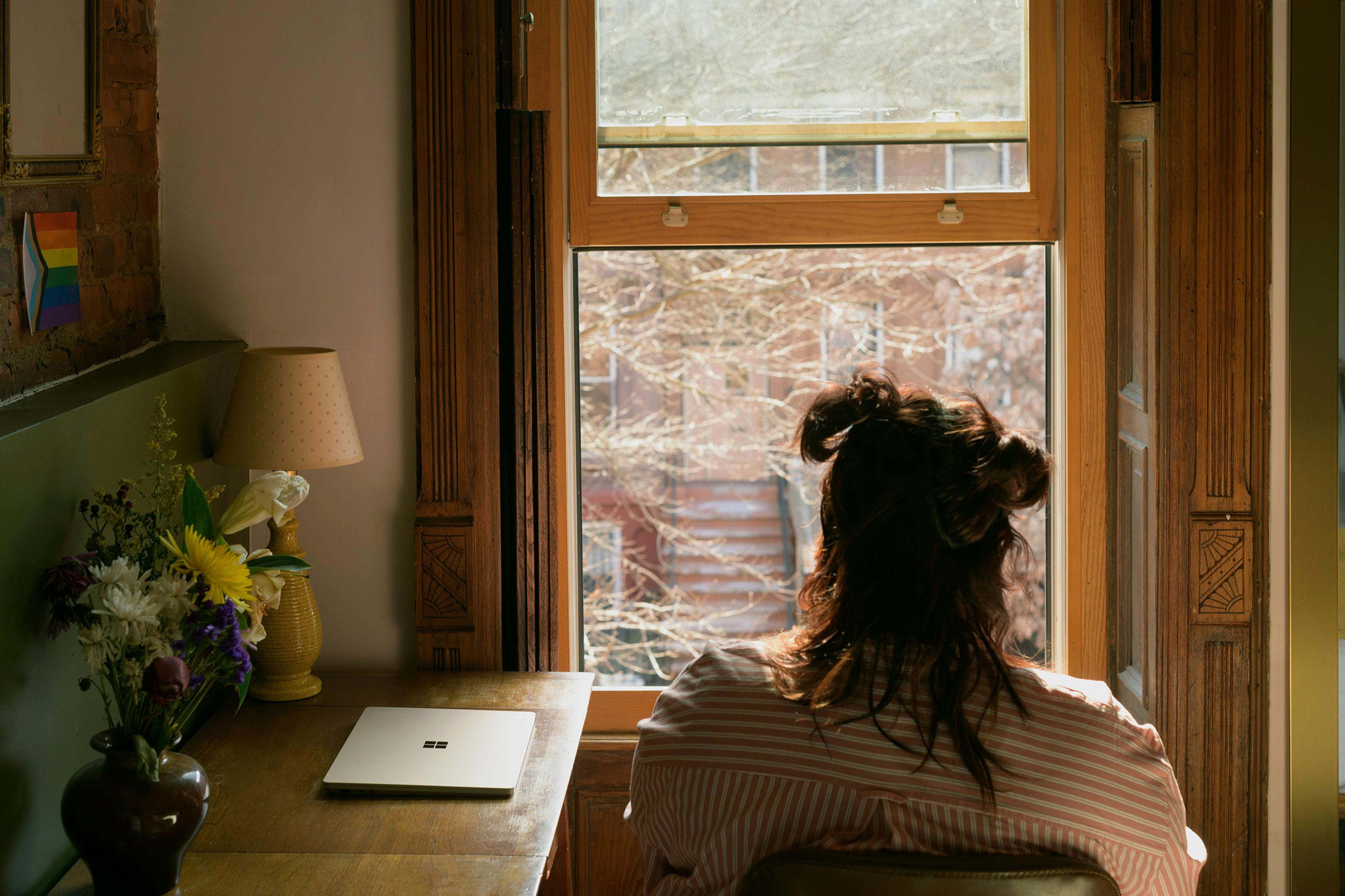 A person with hair up sitting at a desk facing a window, laptop nearby, with flowers and a lamp on the table.