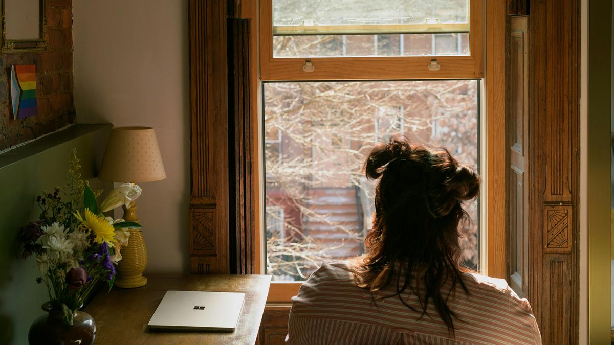A person with hair up sitting at a desk facing a window, laptop nearby, with flowers and a lamp on the table.