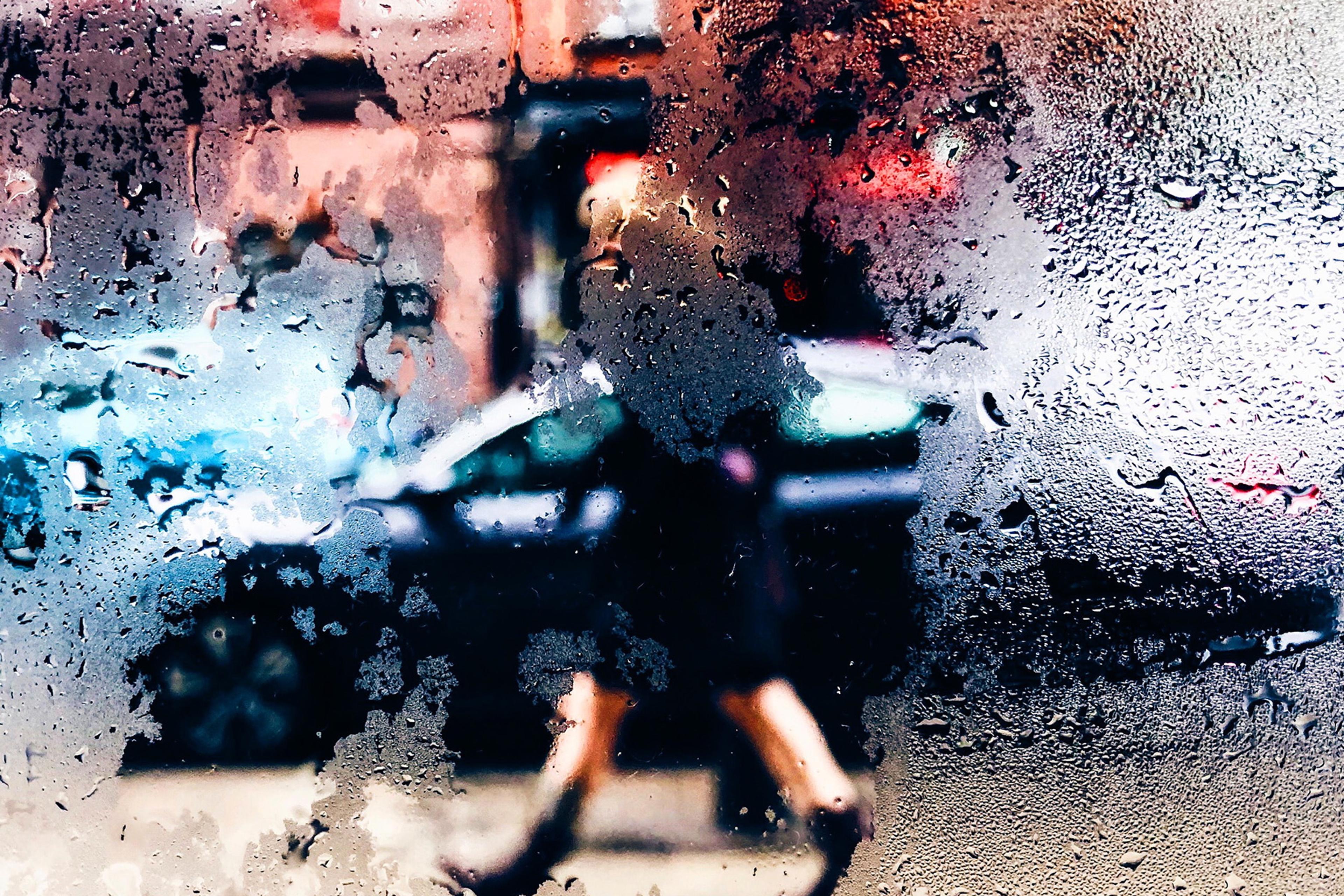 Photo of a blurred street scene viewed through a rainy window, focusing on a pedestrian and coloured buildings in the background.