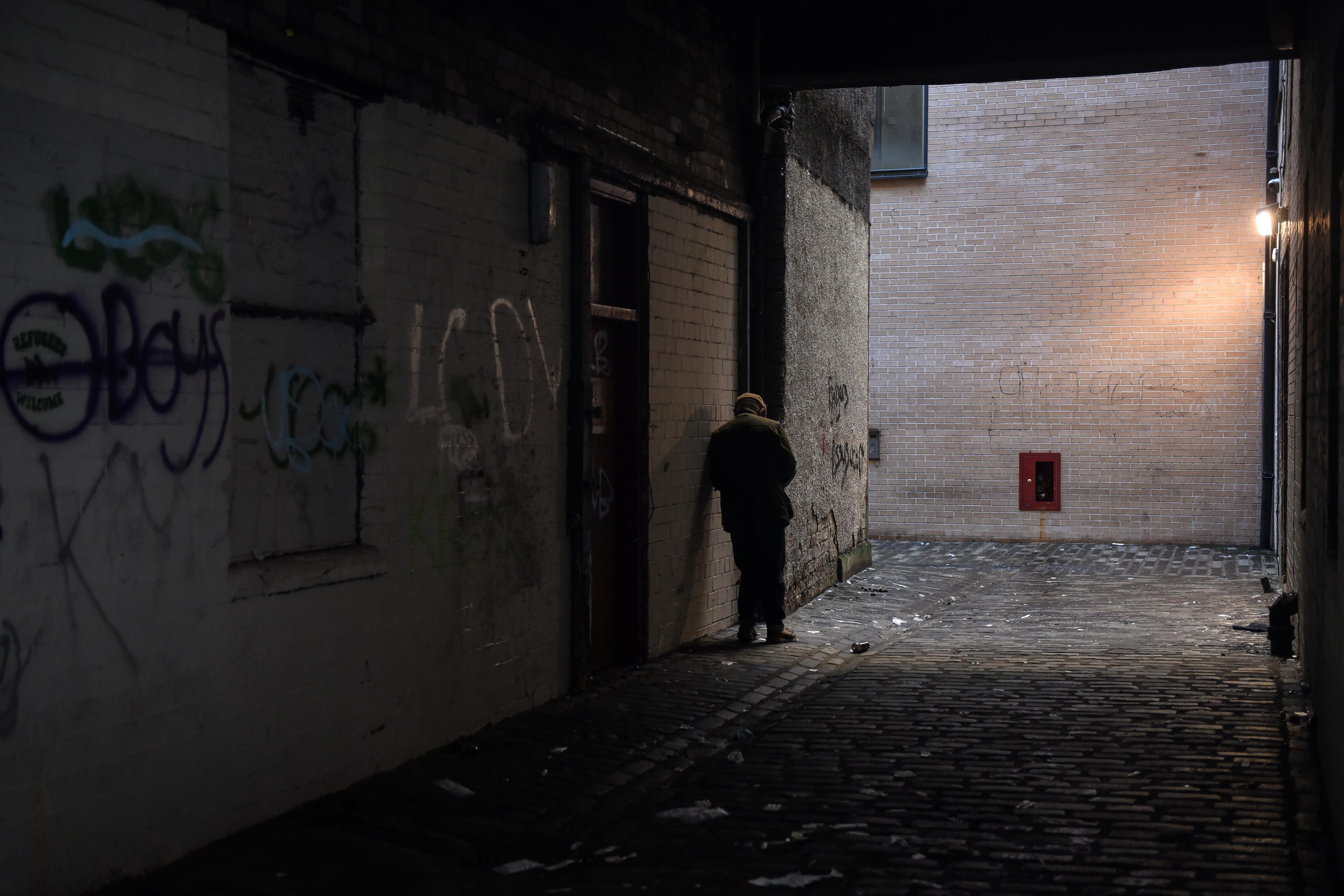 A person leaning against a wall under a light in a dimly lit alley with graffiti-covered walls.