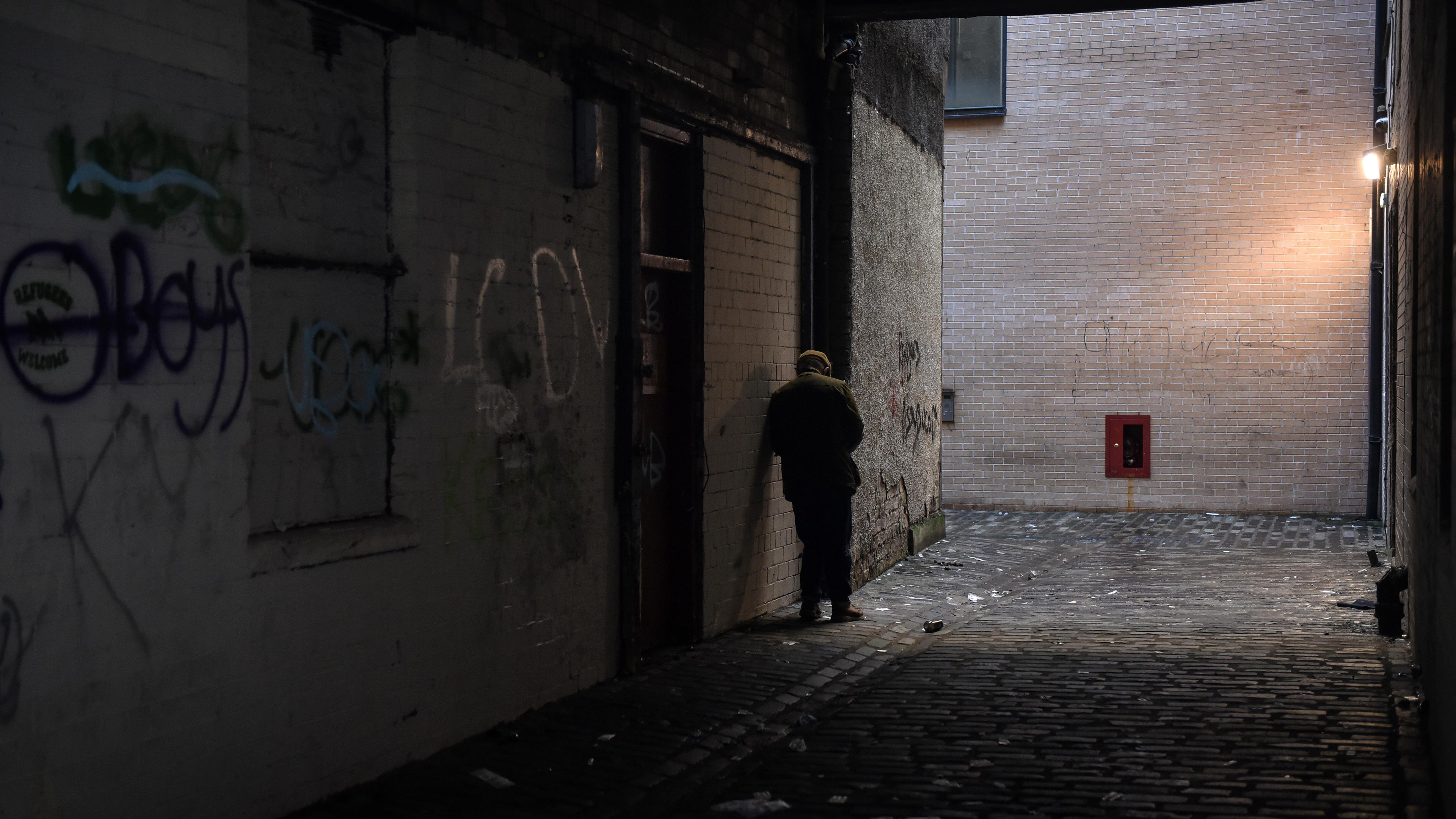 A person leaning against a wall under a light in a dimly lit alley with graffiti-covered walls.