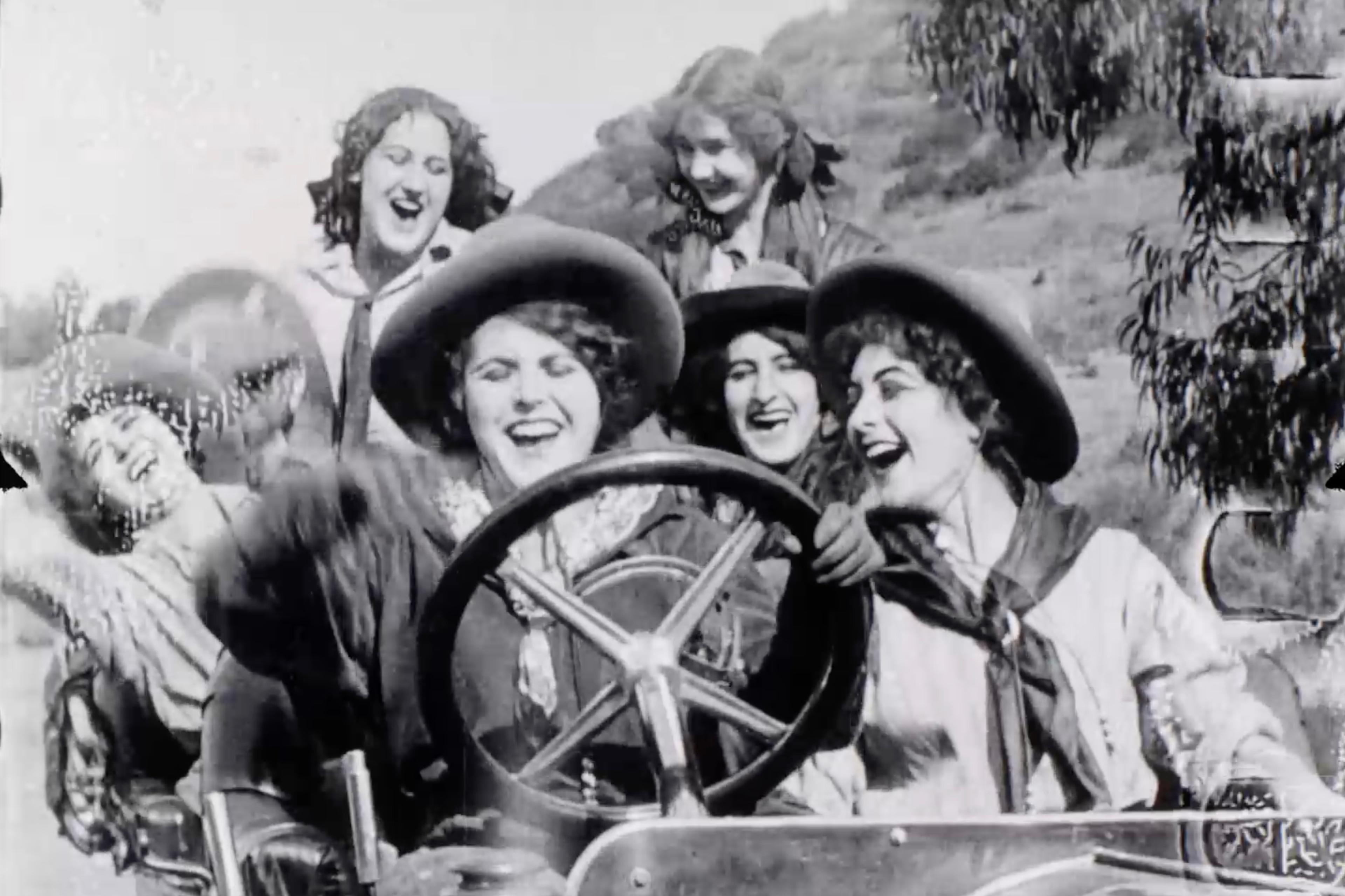 Black and white photo of five women joyfully driving an open car wearing hats surrounded by trees and hills.