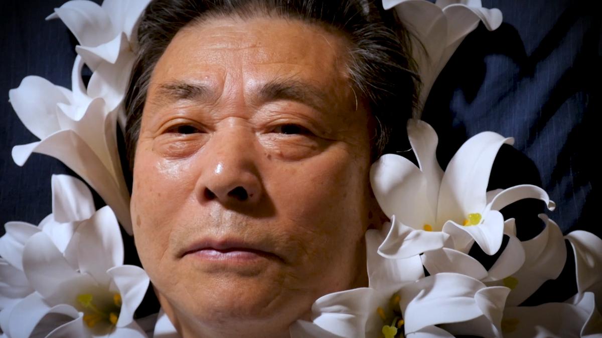A man lying with a serene expression surrounded by white lilies against a dark background.