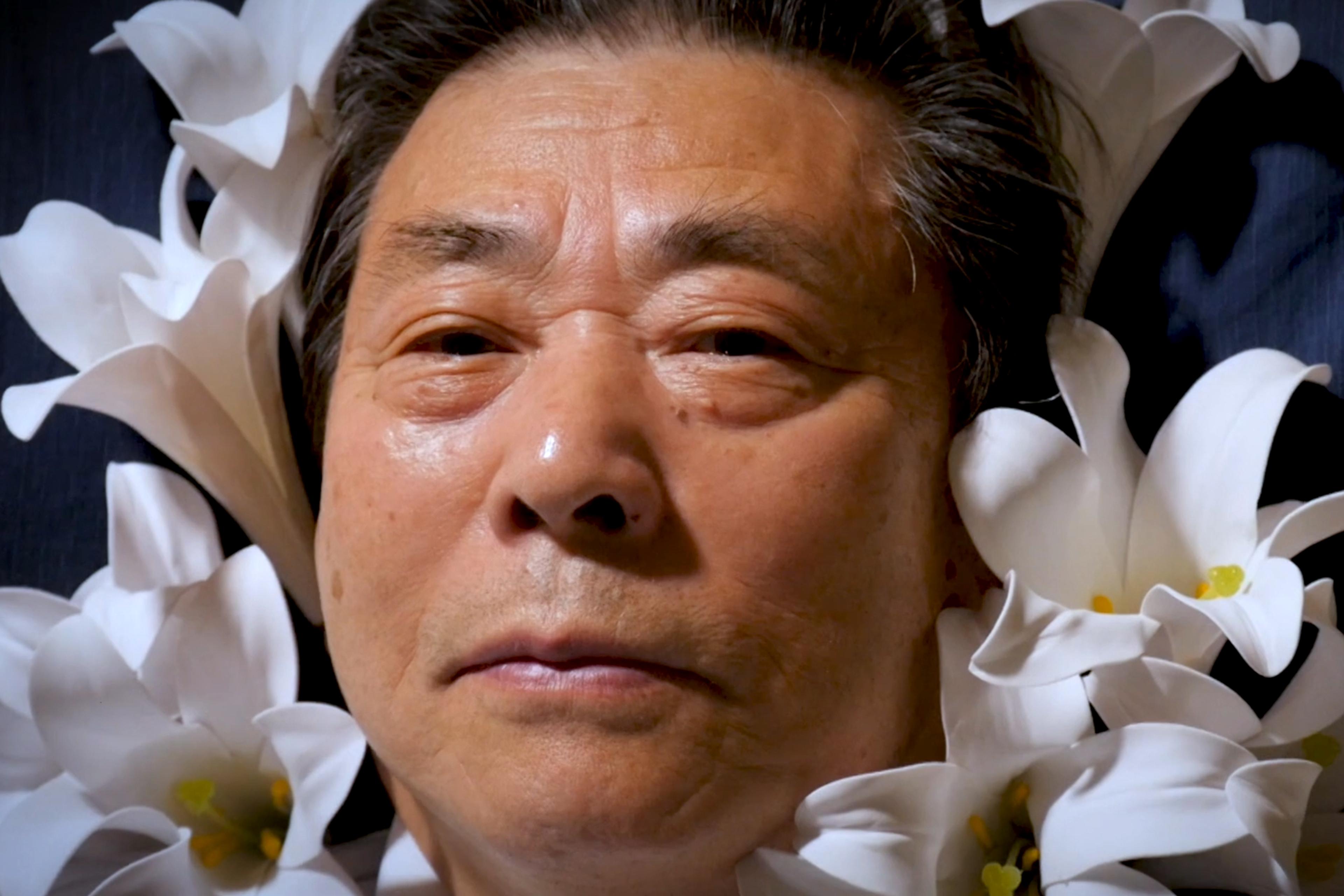 A man lying with a serene expression surrounded by white lilies against a dark background.