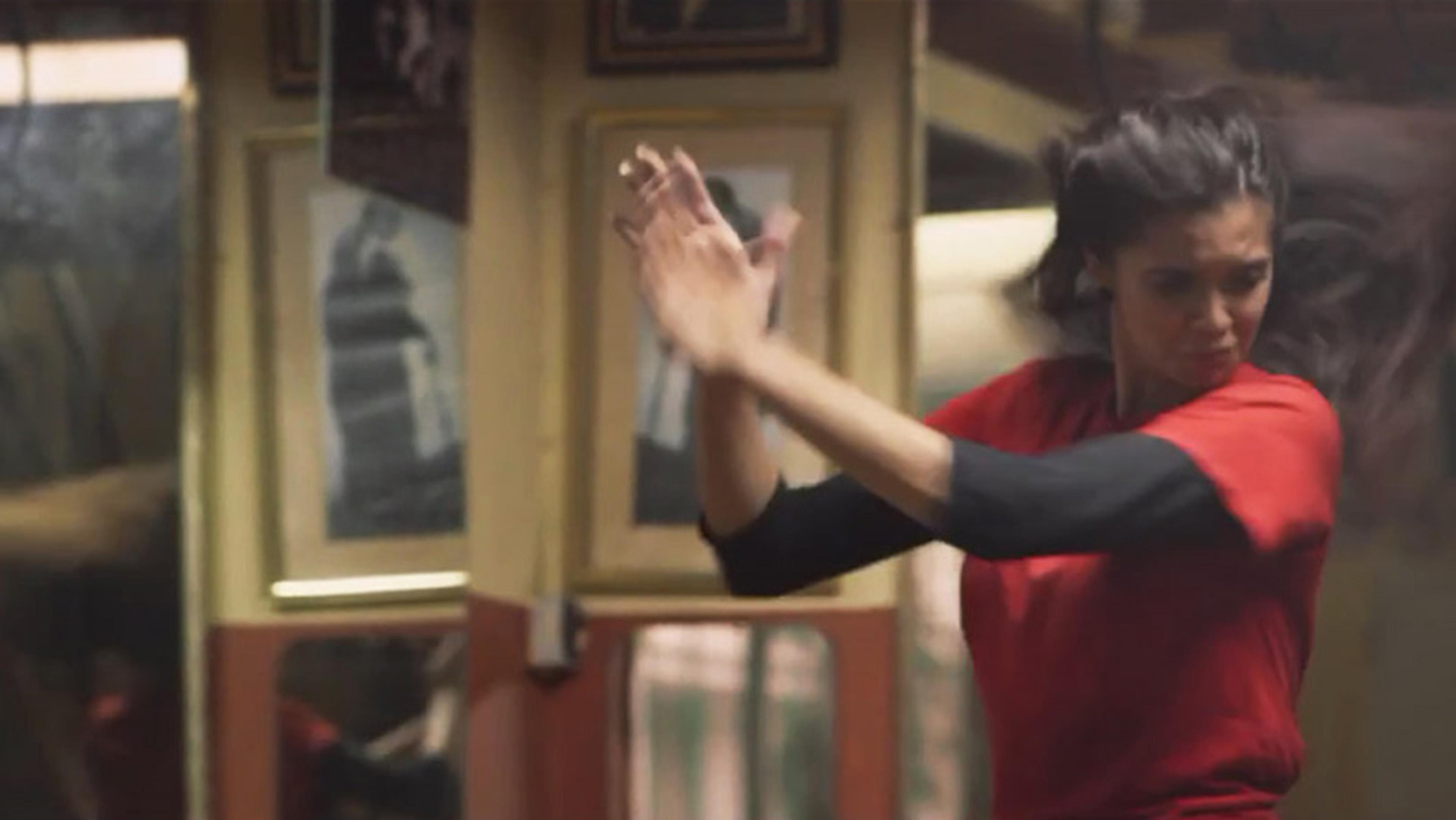 A woman in a red top clapping her hands energetically. She is in mid-motion with blurred movement, surrounded by framed pictures.