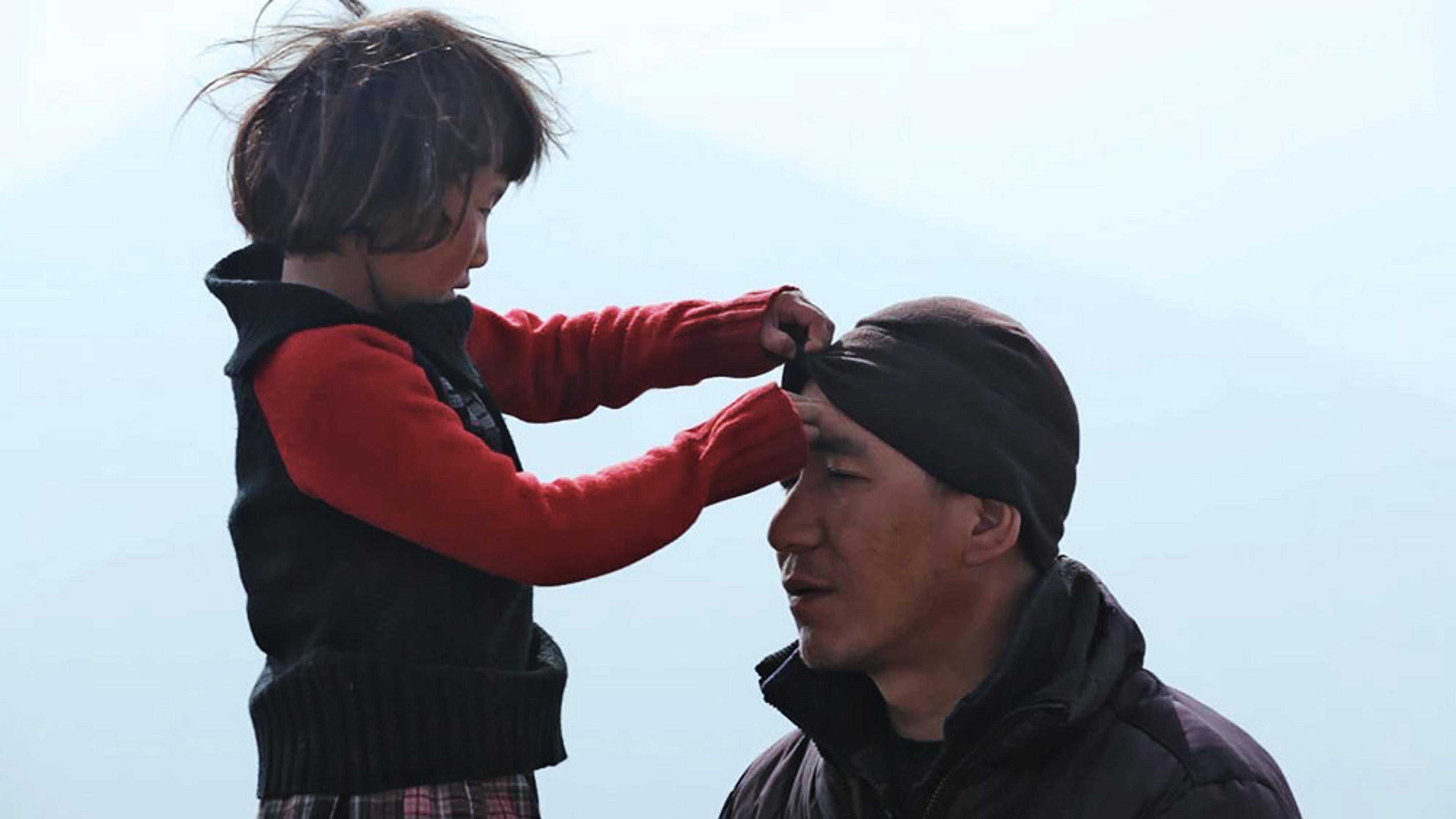 A girl adjusting a man’s hat. The girl is standing while the man is sitting. Both are dressed warmly against a light sky.