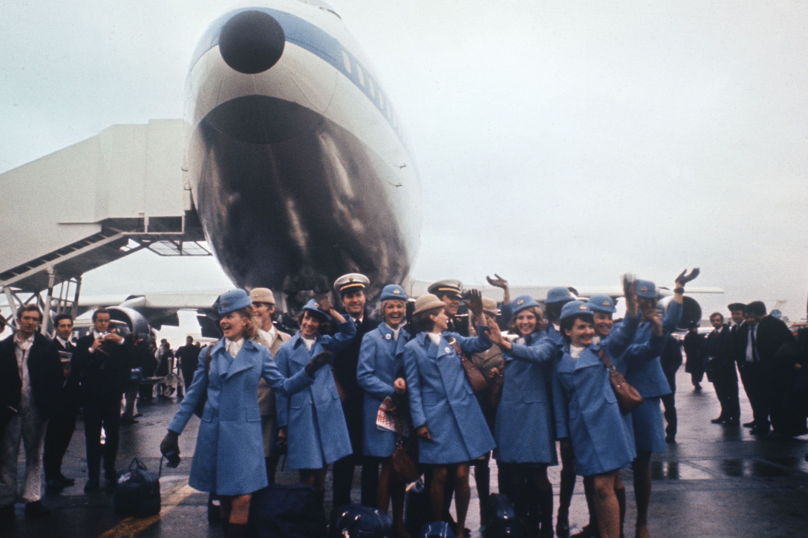 Photo of flight crew in blue uniforms waving in front of a parked aeroplane on a runway, with people in the background.