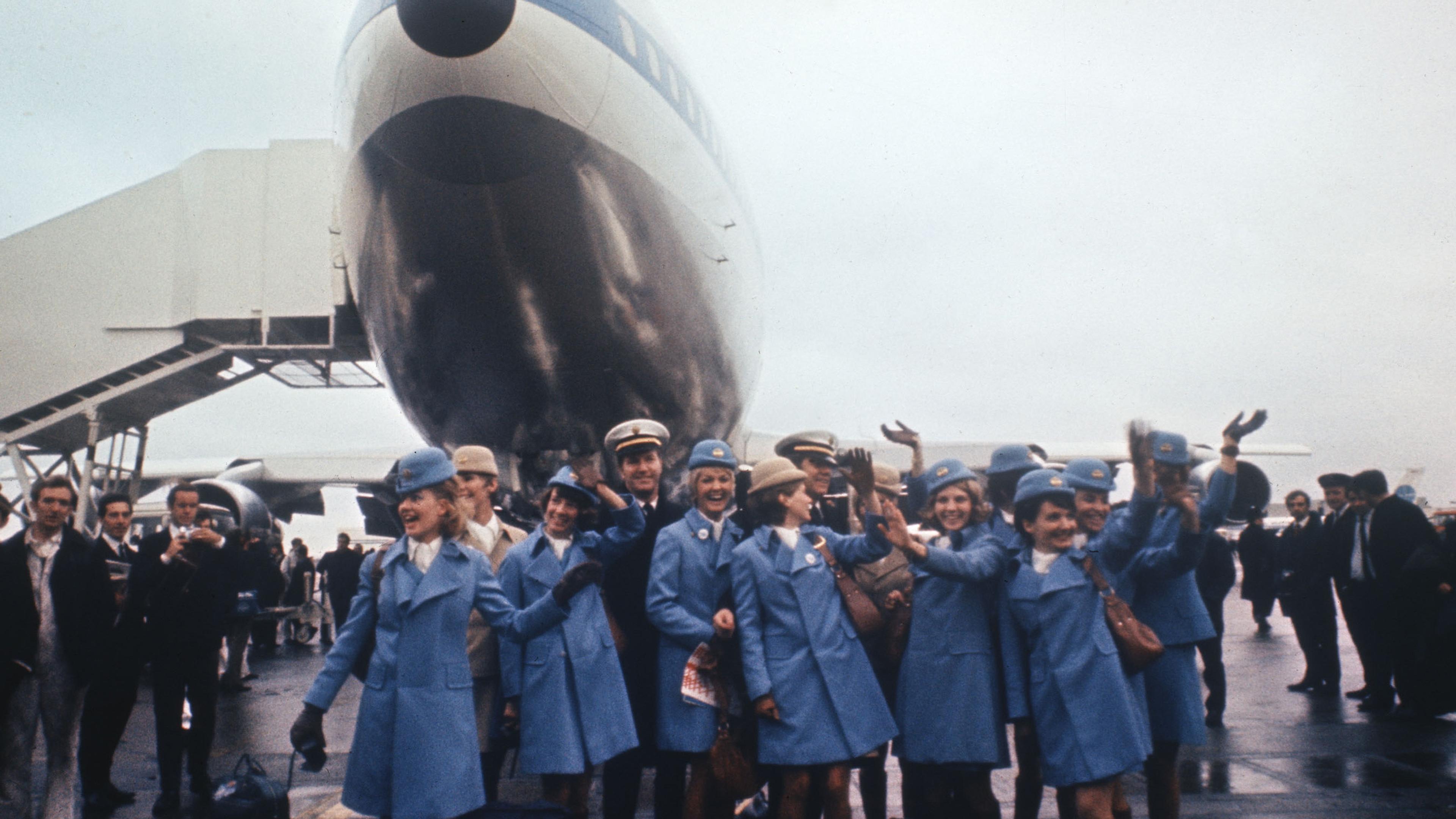 Photo of flight crew in blue uniforms waving in front of a parked aeroplane on a runway, with people in the background.