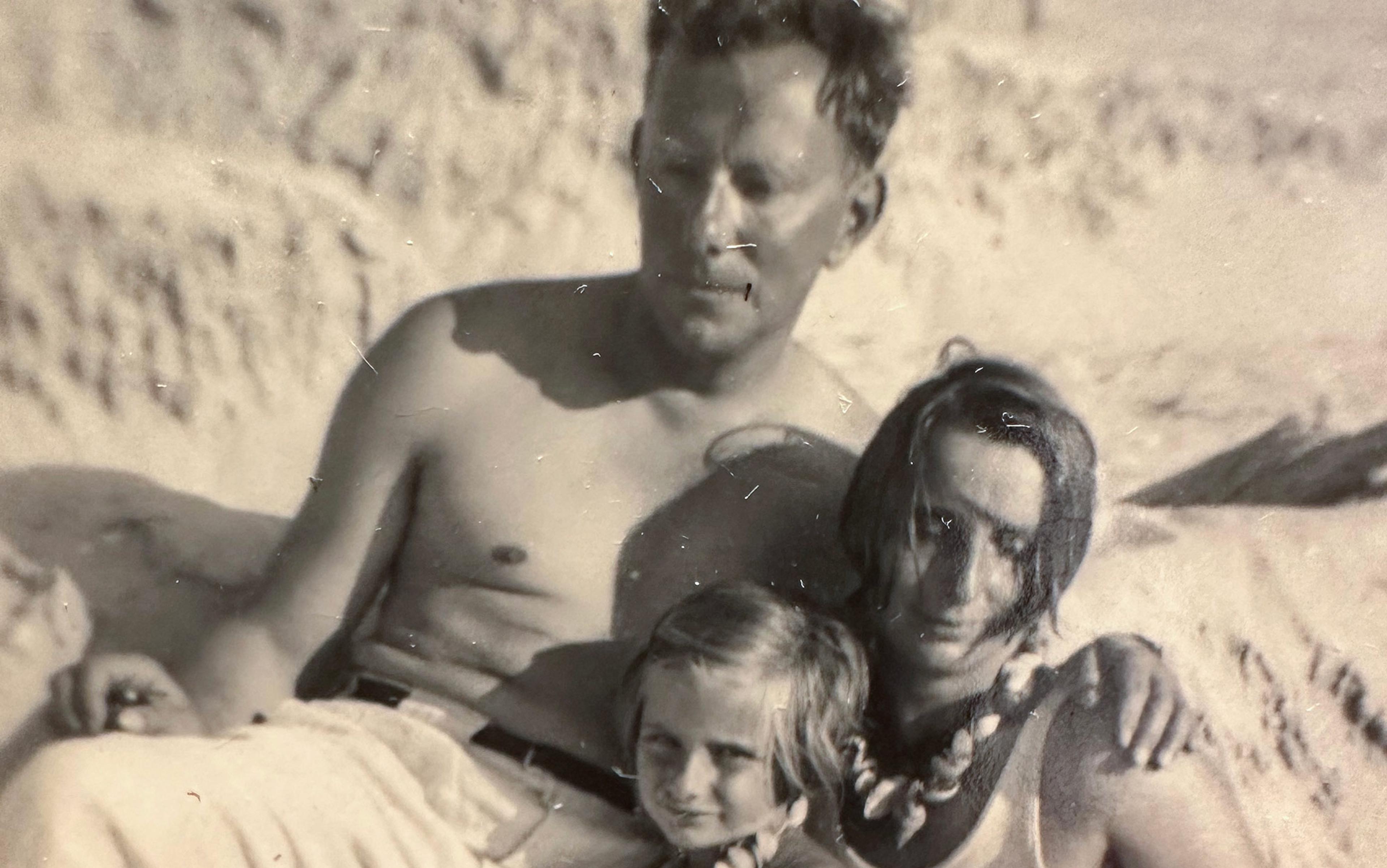 A sepia photo of a shirtless man, a woman, and a child sitting on a beach; the child and woman wear shell necklaces.