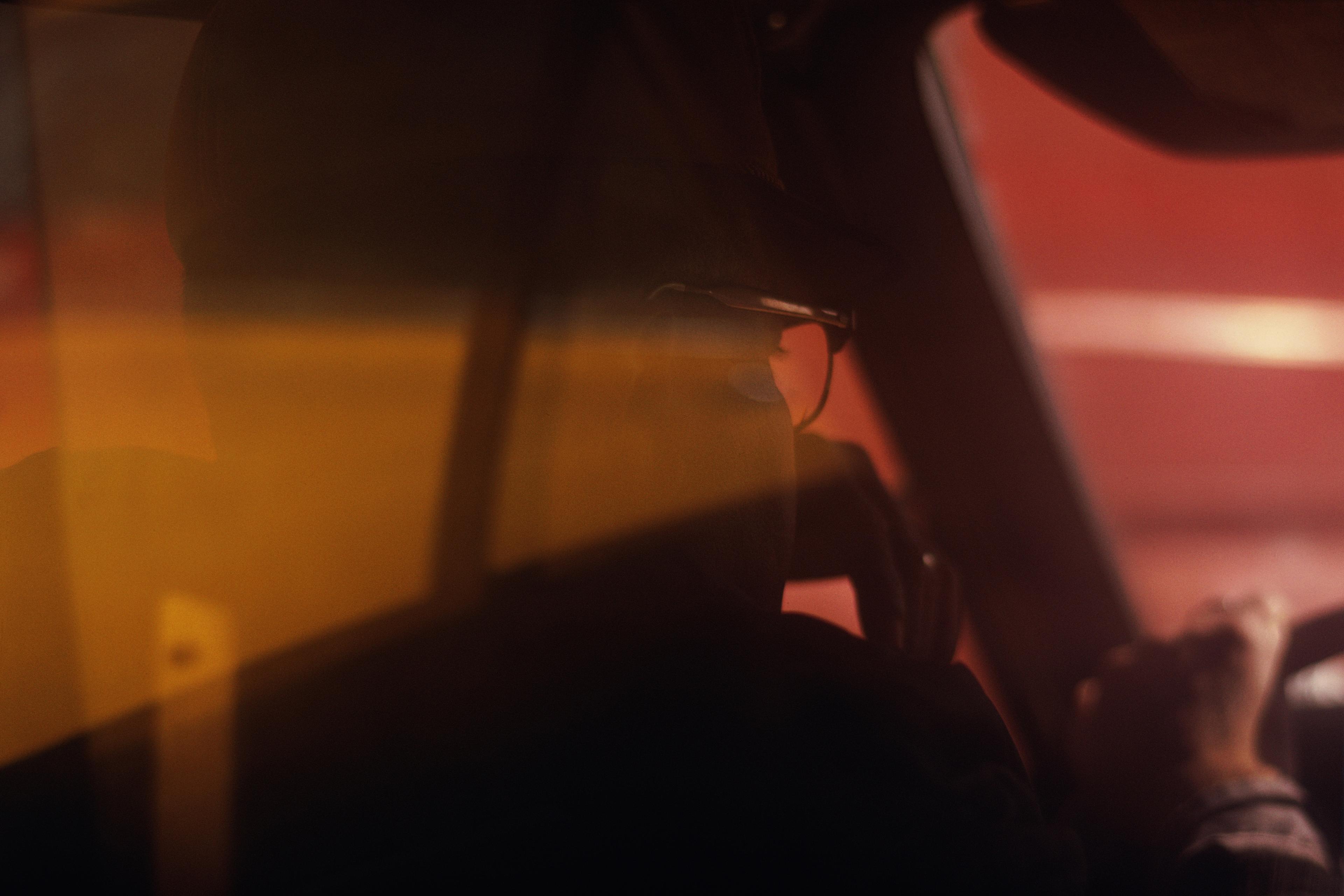 Photo of a person driving a car, seen from the back seat with reflections creating an abstract effect.