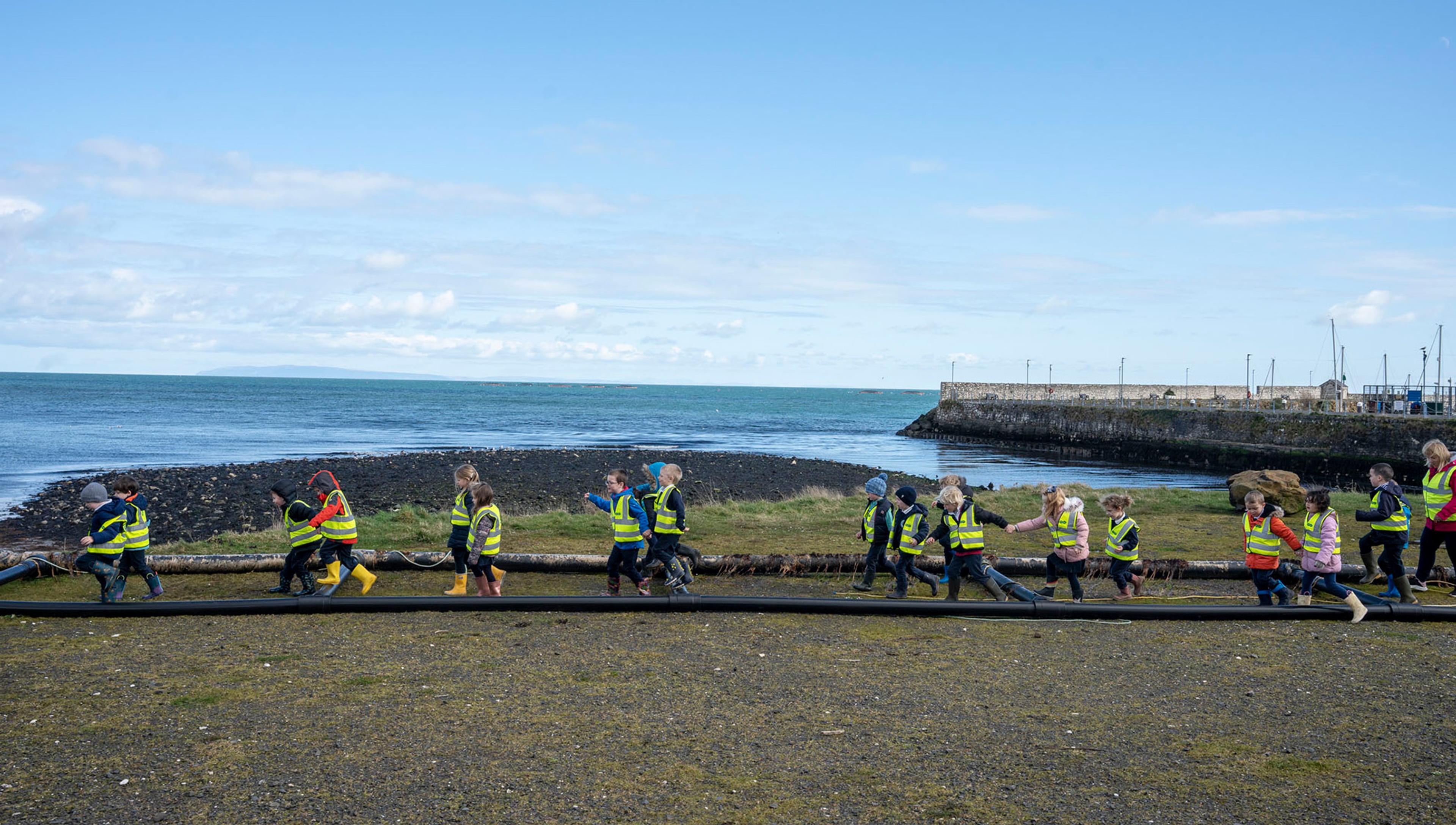 A group of school children in high-vis vests walk alongside a harbour wall beneath a blue sky