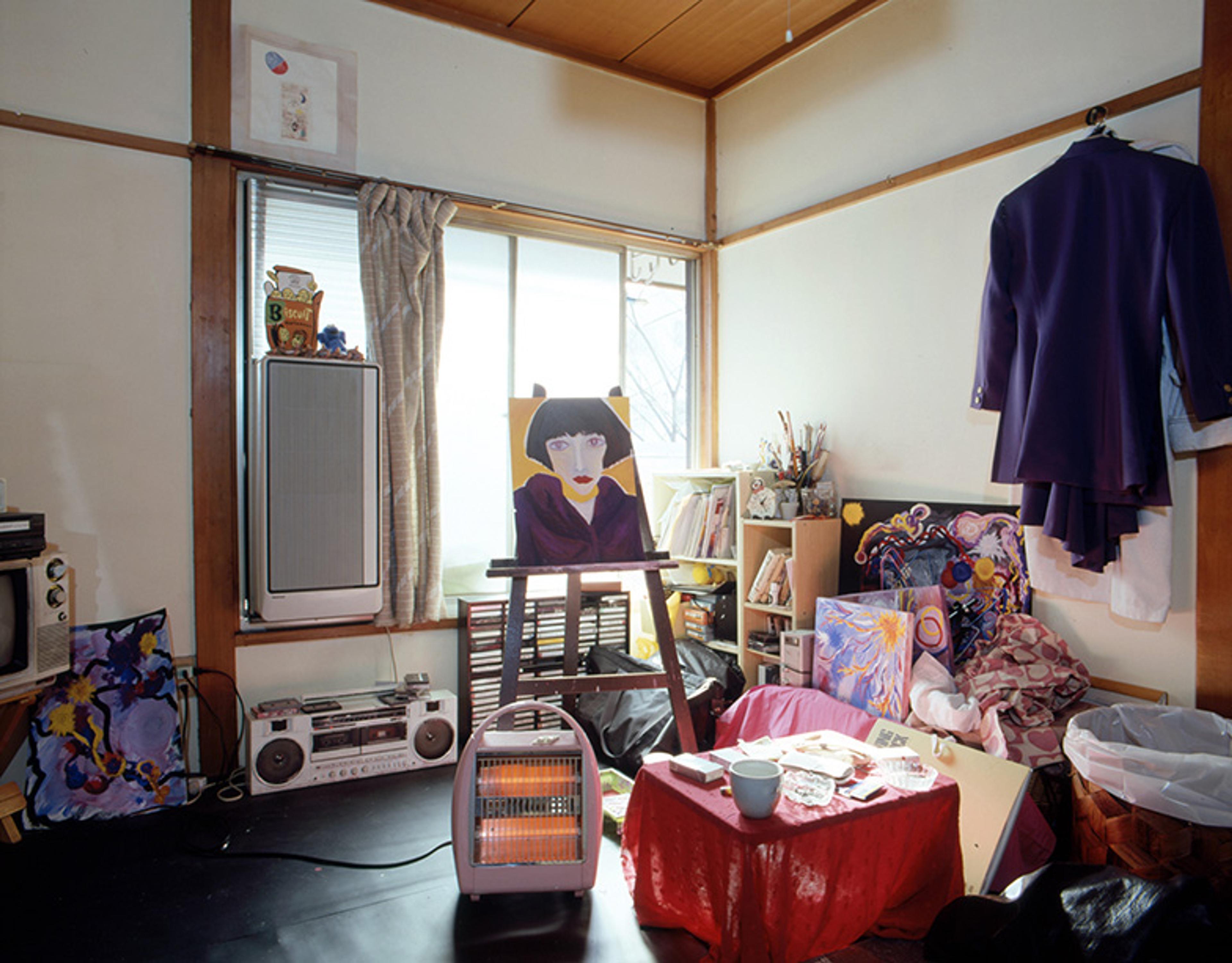 A cluttered room with an easel displaying a portrait, a stereo, books, a purple suit hanging, and various painted canvases and art supplies scattered around.