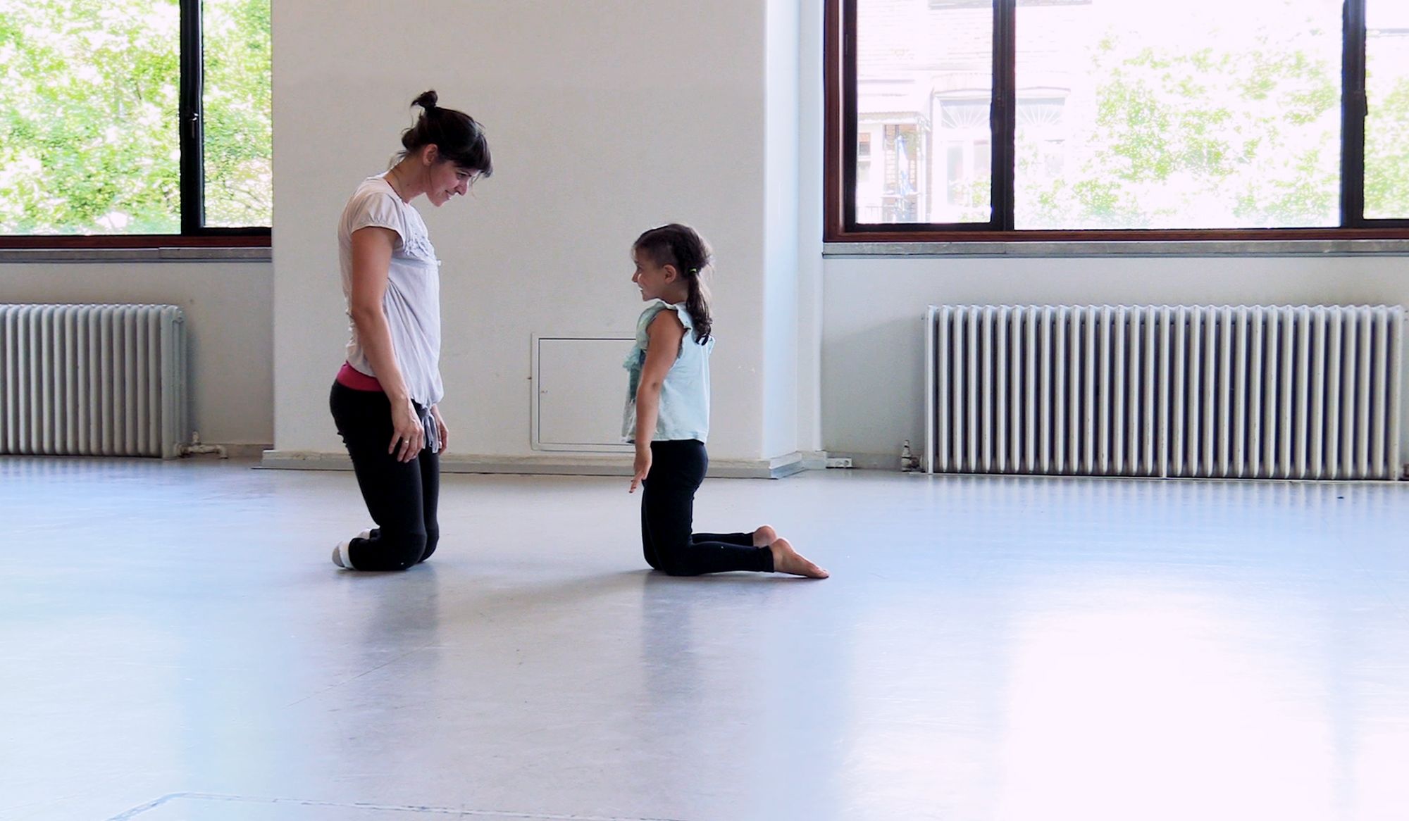 An adult and a child kneeling opposite each other on a studio floor with large windows and radiators in the background.