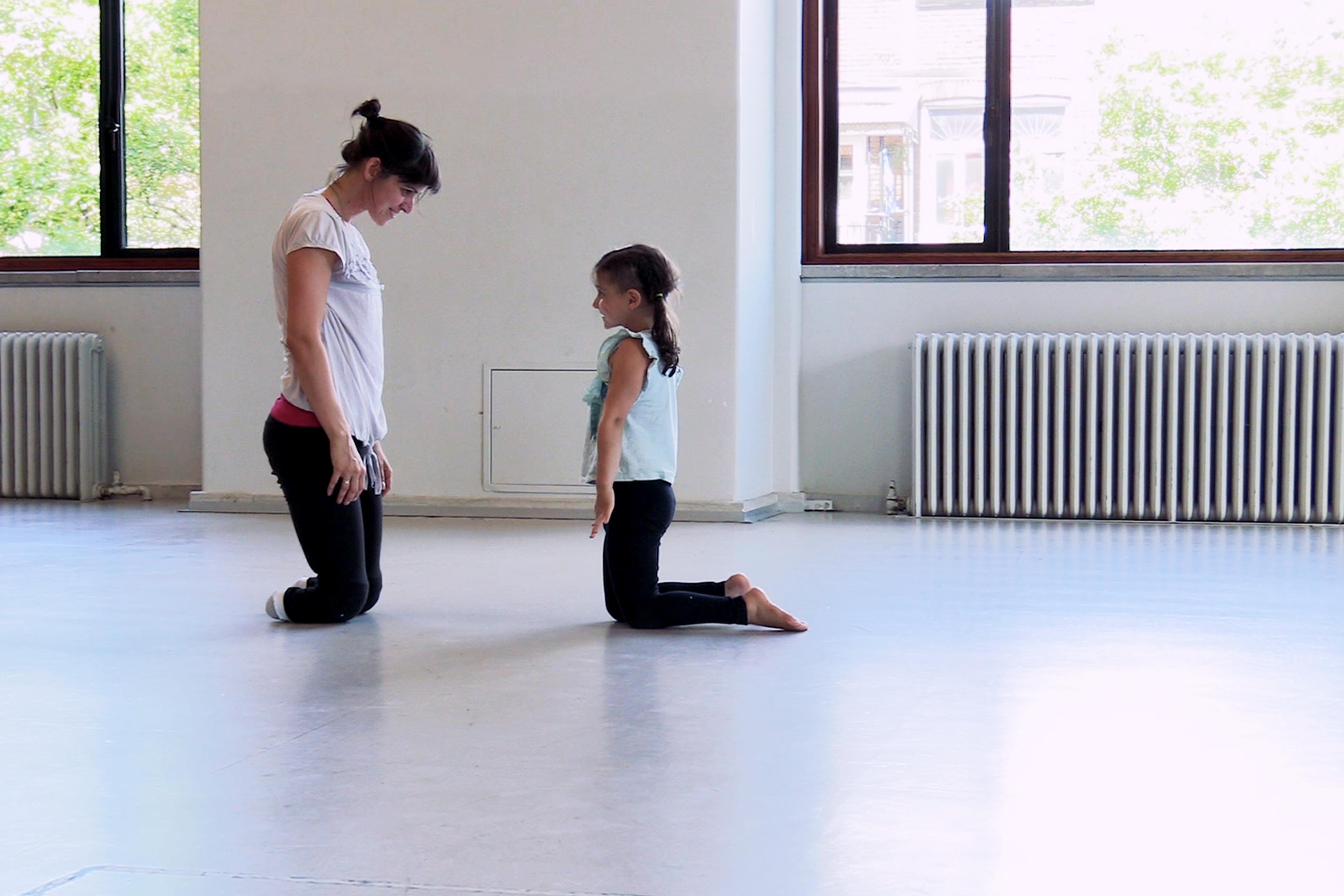 An adult and a child kneeling opposite each other on a studio floor with large windows and radiators in the background.